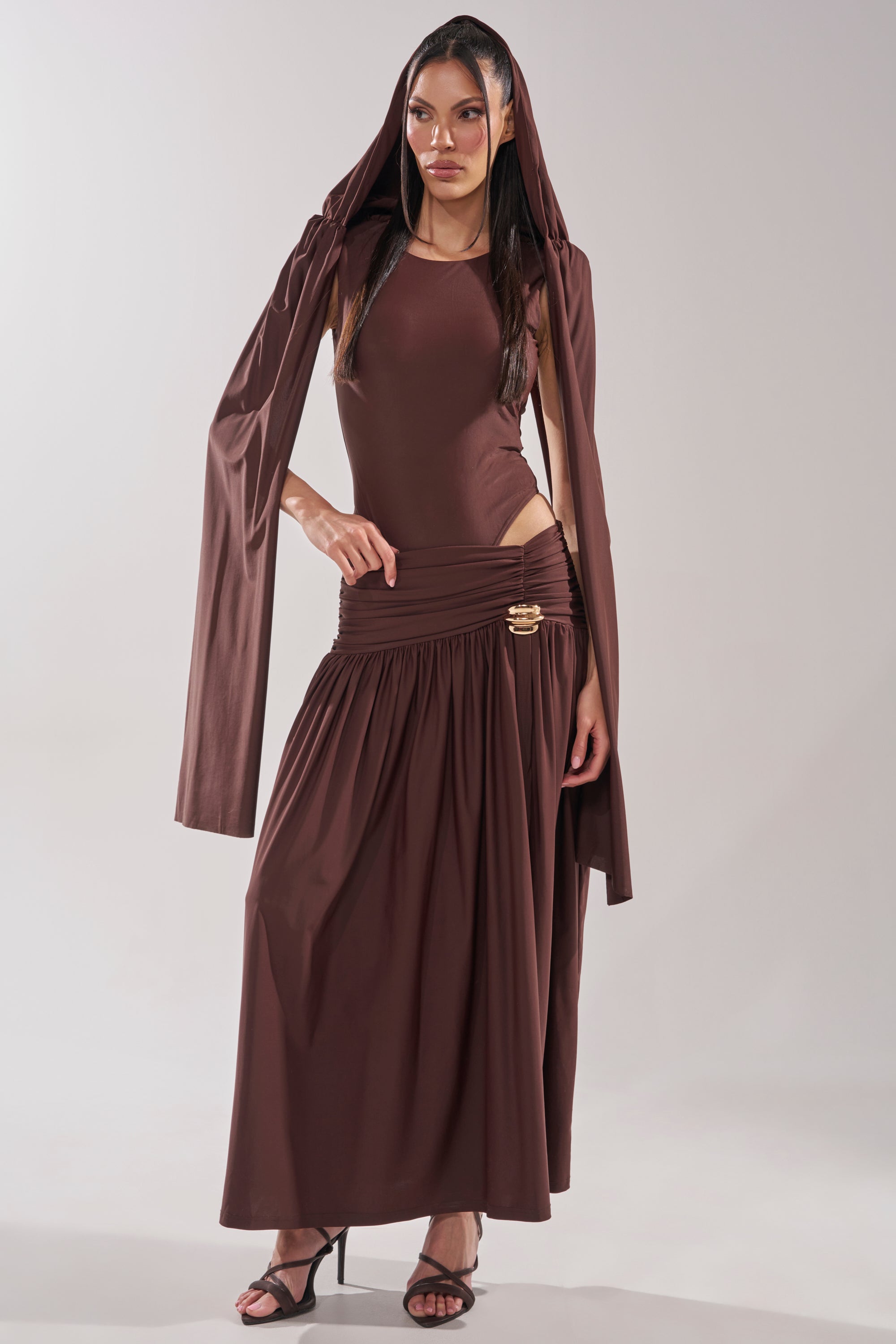 A woman stands confidently in heels wearing the HILLARY SEAMLESS BODYSUIT, a long brown dress with draped sleeves, head covering, gathered hips, gold clasps, and set against a plain light background. 10