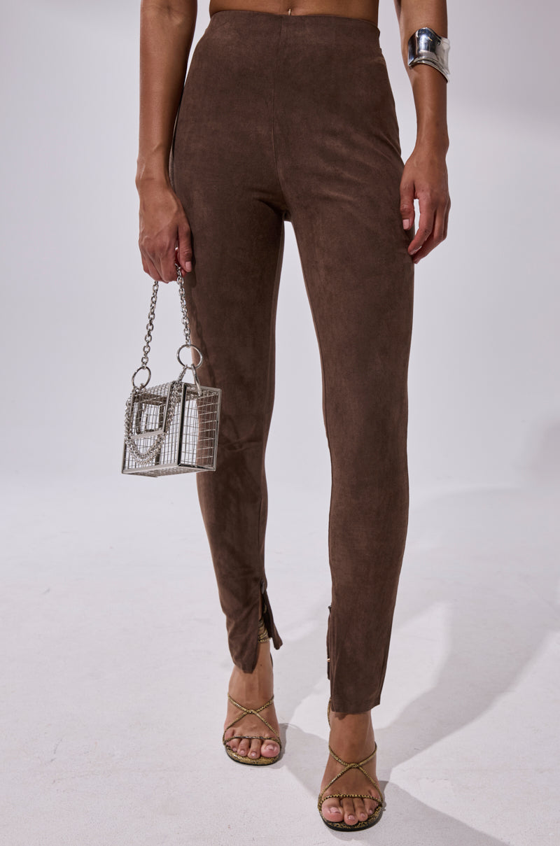 Someone wears CHOCOLATE HEAVEN SUEDE LEGGING with ankle zippers, strappy sandals, a metallic cuff, and holds a small silver chain purse. The background is plain and light-colored.