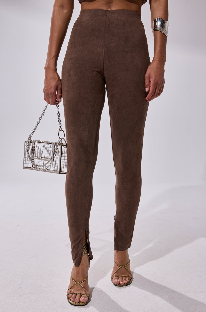 A woman stands against a plain light background wearing CHOCOLATE HEAVEN SUEDE LEGGING, strappy heeled sandals, a silver cuff bracelet, and holding a small metal cage-style handbag.
