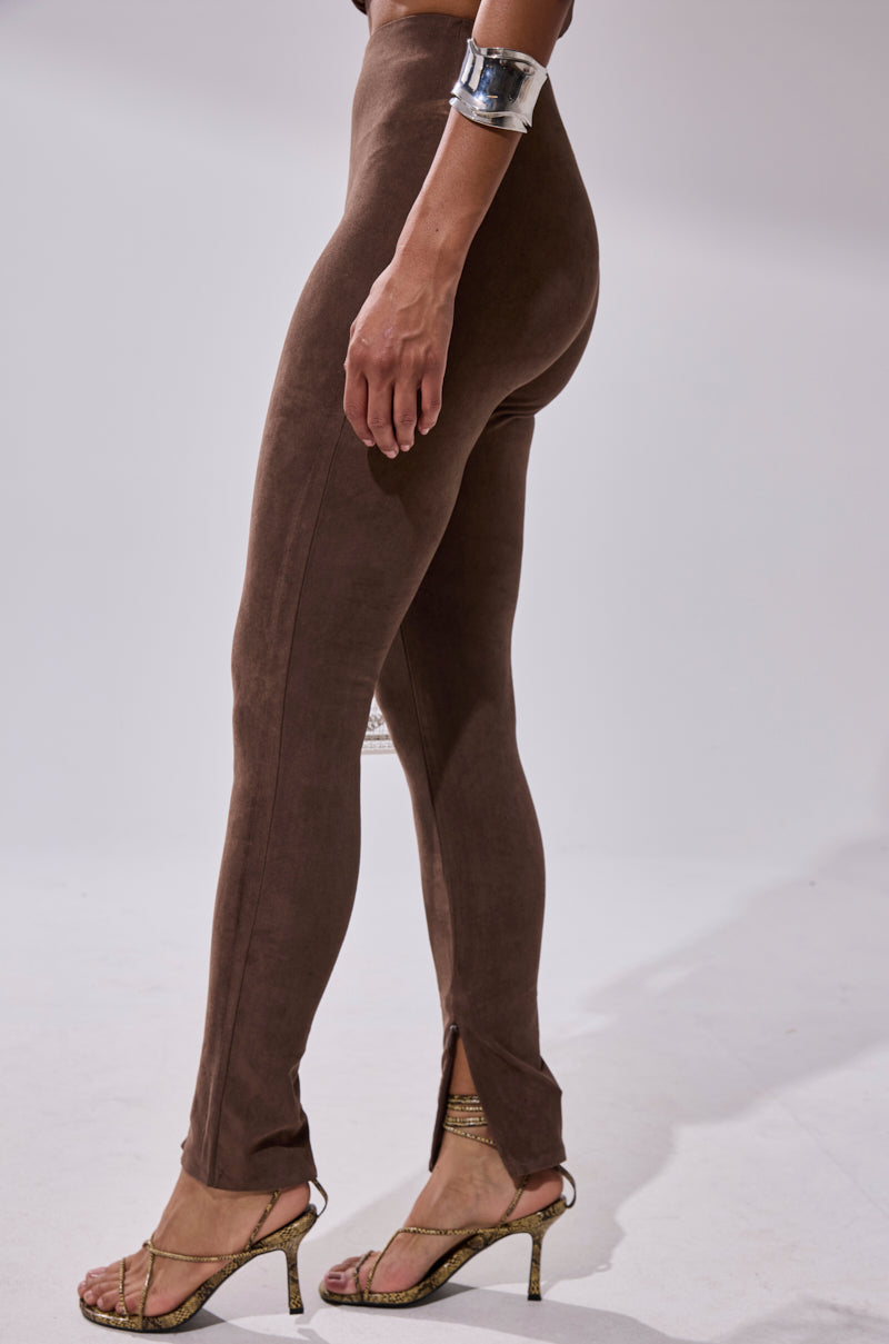 A person wearing CHOCOLATE HEAVEN SUEDE LEGGING with front ankle slits, metallic heeled sandals, and a large silver cuff bracelet stands against a plain light background.