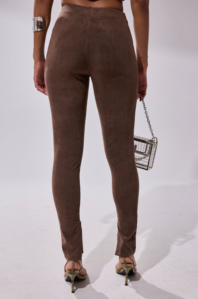 A person faces away, wearing CHOCOLATE HEAVEN SUEDE LEGGING, high-heeled sandals, a chunky silver bracelet, and carrying a small metallic handbag against a plain light background.
