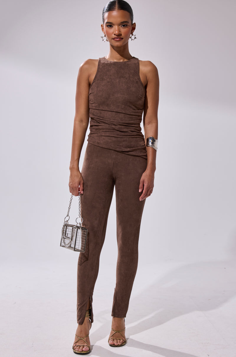A woman stands facing forward in a sleeveless, fitted brown top and CHOCOLATE HEAVEN SUEDE LEGGING. She carries a small handbag, wears heeled sandals, hoop earrings, a cuff bracelet, and her hair is styled in a sleek bun.