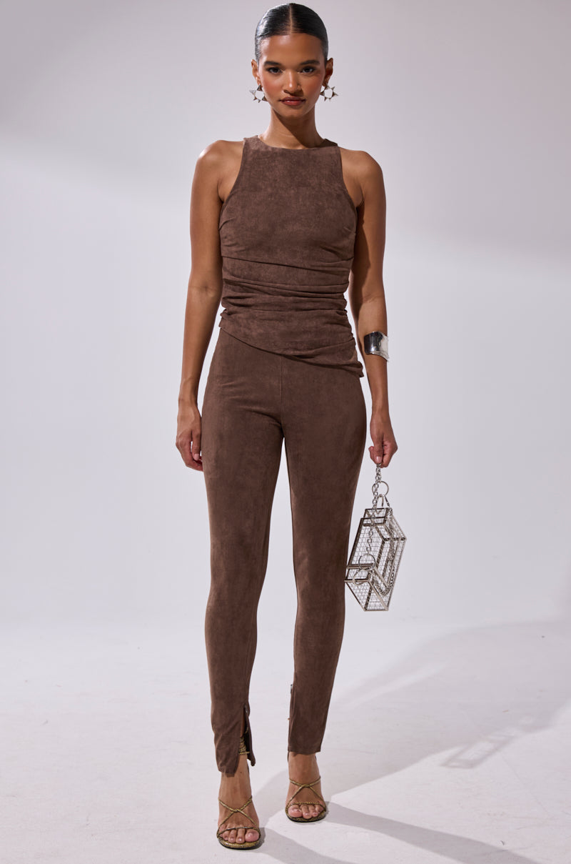 A woman poses against a plain backdrop wearing the CHOCOLATE HEAVEN SUEDE BLOUSE and matching pants, highlighting a flattering silhouette. She completes the look with statement earrings, a cuff bracelet, strappy heels, and a metallic handbag.