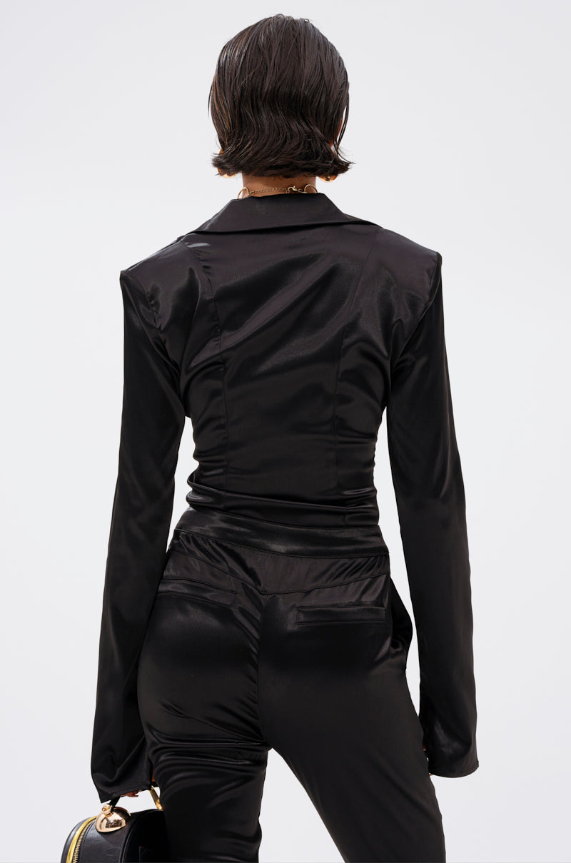 A person with short dark hair is shown from behind wearing the DOWN FOR WHATEVER BUTTON DOWN BODYSUIT under a fitted black satin blazer and pants, holding a black bag with a gold handle, against a plain light background. 3