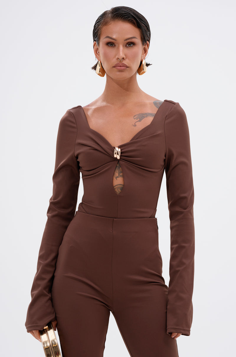 A person with short dark hair wears the LUXE LIFE SEAMLESS BODYSUIT IN BROWN, a curve-sculpting long-sleeve jumpsuit with a front cutout and gold clasp. They accessorize with gold geometric earrings, a gold clutch, and display a shoulder tattoo against a white background.