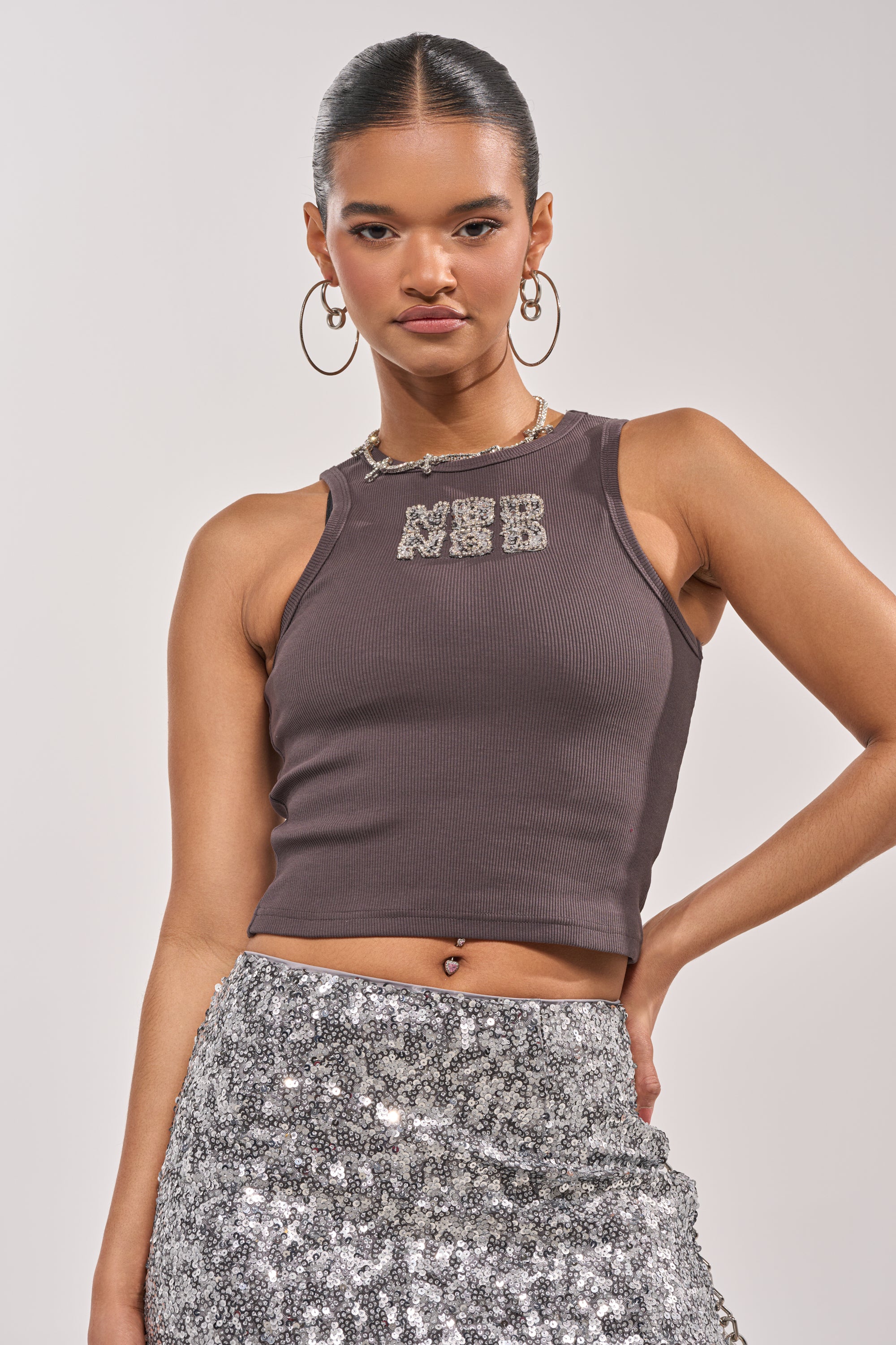 A woman poses with one hand on her hip, wearing the NO BIG DEAL TANK—a dark ribbed style with rhinestone lettering—paired with a silver sequin skirt, hoop earrings, a necklace, and her hair pulled back for a Y2K-inspired look. 0