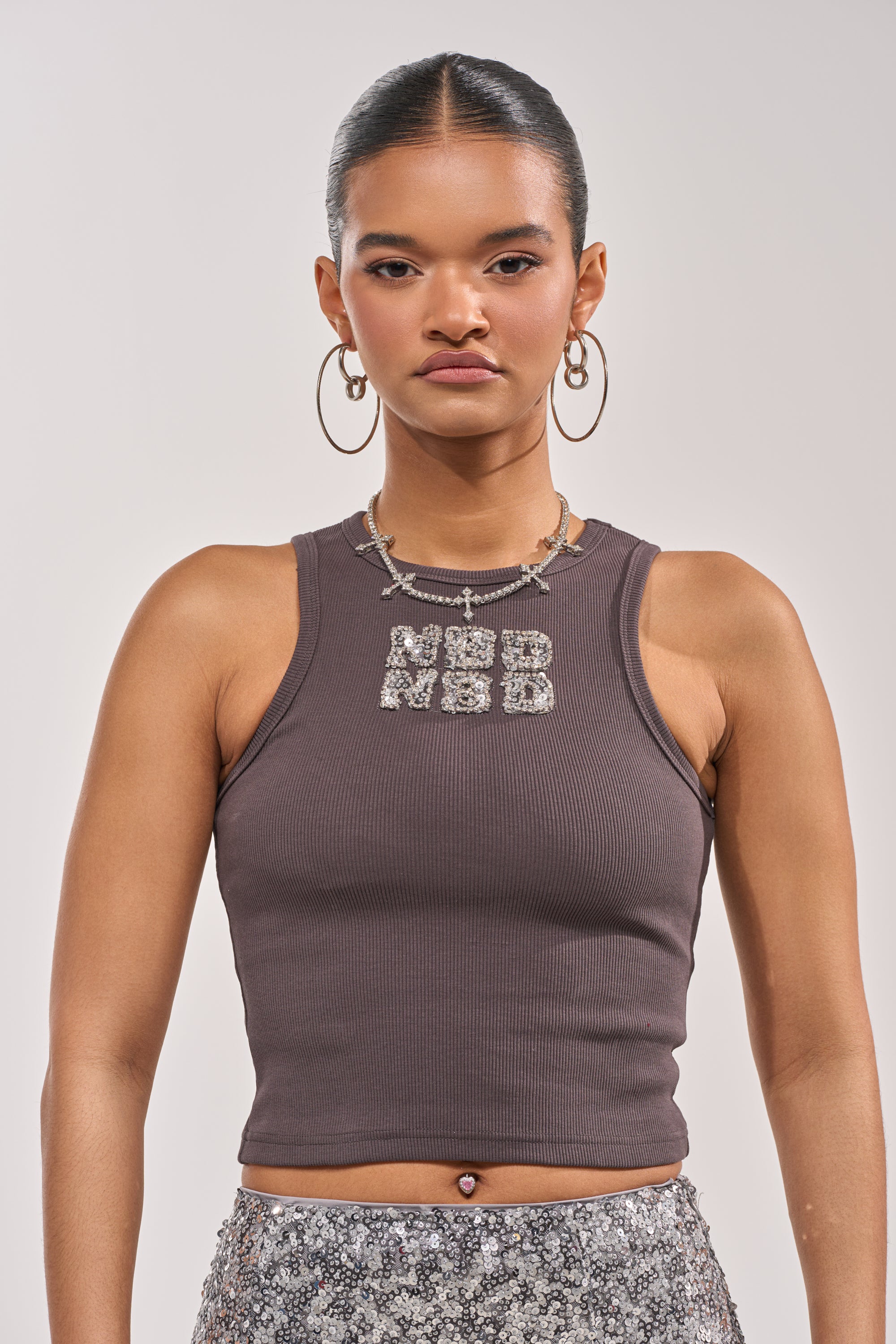 A woman with slicked-back hair wears large hoop earrings, layered necklaces—one with sequin lettering—and the Y2K-inspired NO BIG DEAL TANK. She stands against a plain light background. 1