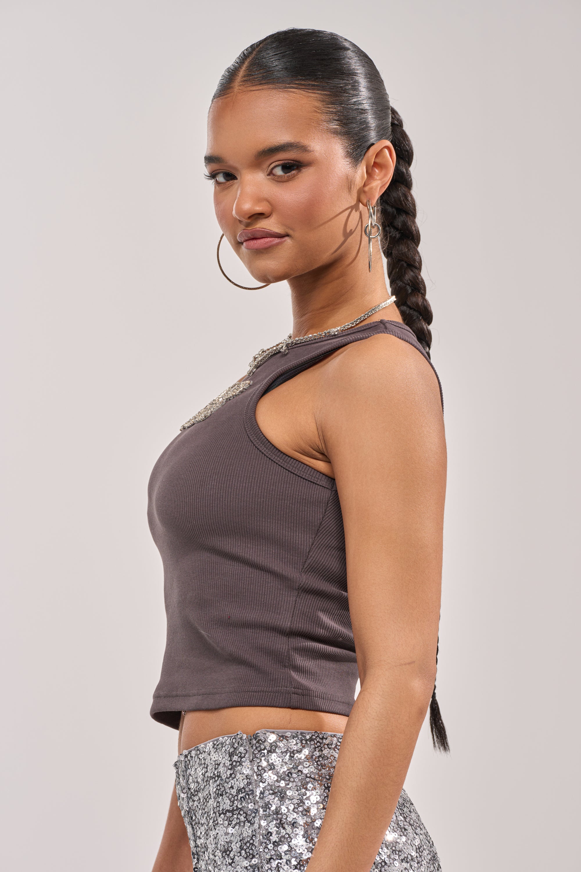 A woman with braided hair and hoop earrings wears the NO BIG DEAL TANK in brown with shiny sequin pants, channeling Y2K style as she stands sideways, looking confidently at the camera against a plain background. 2