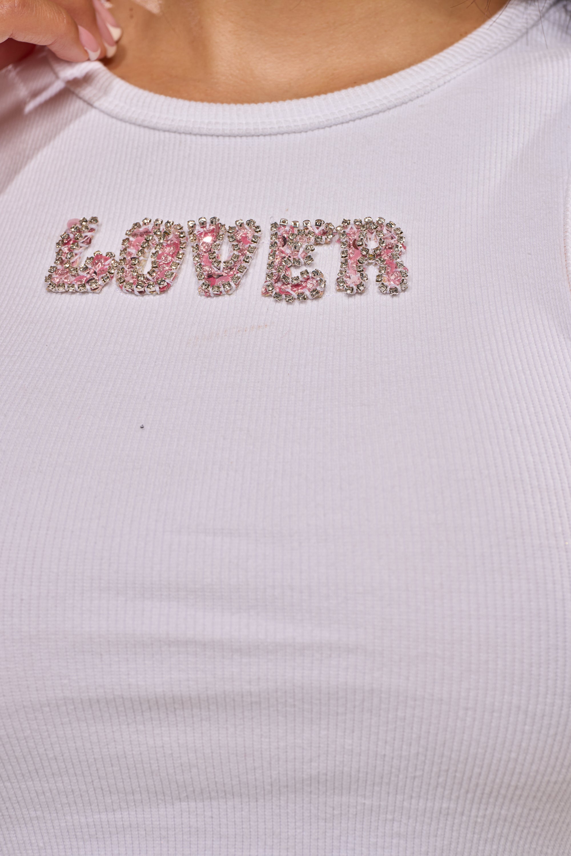 Close-up of the LOVER GIRL RIBBED TANK by AKIRA Label, a white ribbed tank featuring "LOVER" in pink and silver rhinestones across the chest. 10