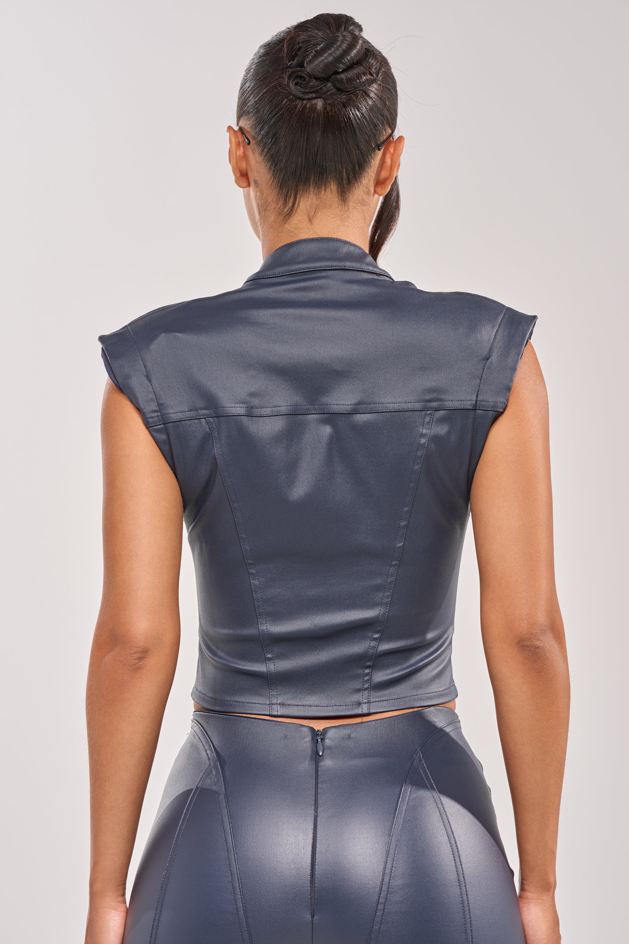 A woman with a twisted updo wears the BIG CITY LIVIN FAUX LEATHER BUTTON DOWN and matching high-waisted pants, styled in a sleek dark faux leather. She is shown from the back against a plain light background. 3