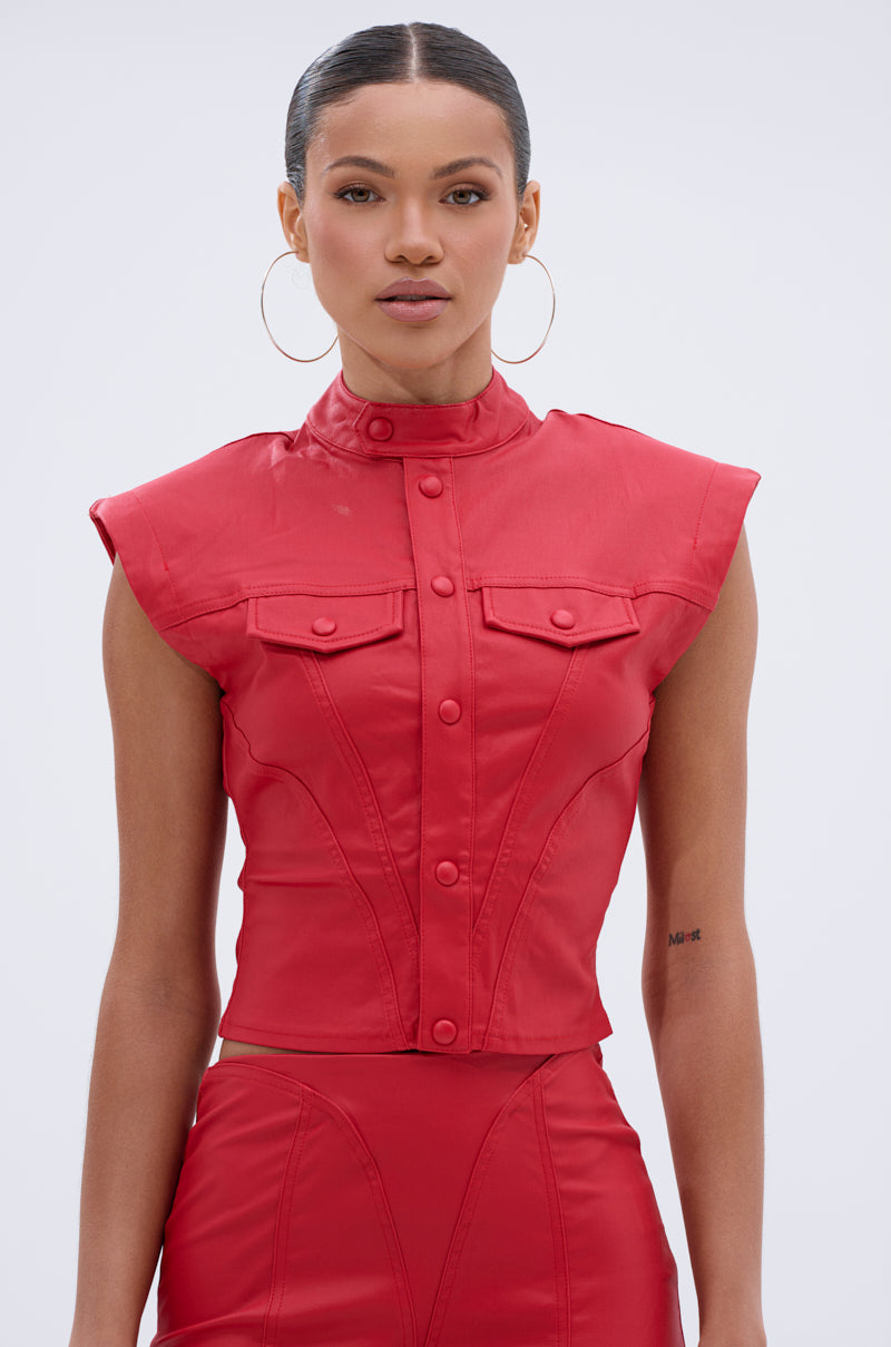 A woman with slicked-back hair wears the BIG CITY LIVIN FAUX LEATHER BUTTON DOWN, a fitted sleeveless red top with front pockets and button details, styled with matching high-waisted bottoms, large hoop earrings, and a small tattoo on her left forearm.