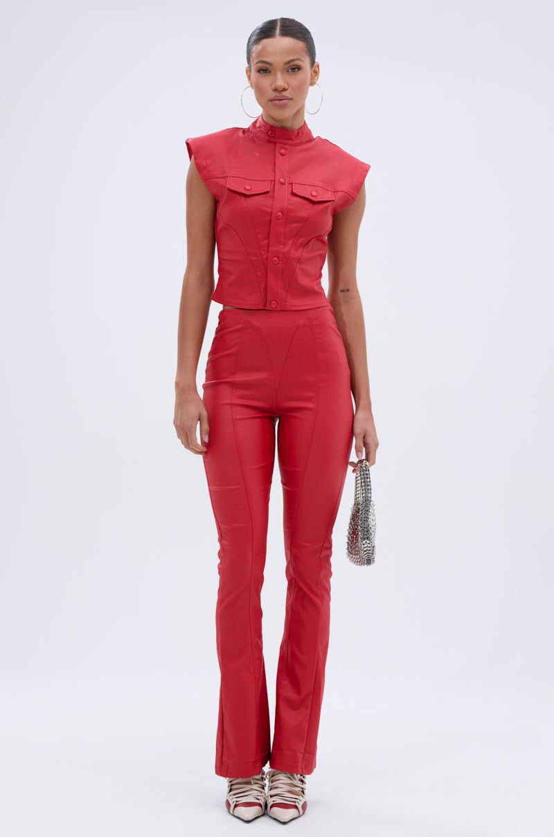A woman poses against a plain backdrop wearing the BIG CITY LIVIN FAUX LEATHER BUTTON DOWN—sleeveless, fitted, with button accents and a moto collar—paired with a small silver handbag, hoop earrings, and patterned heels.