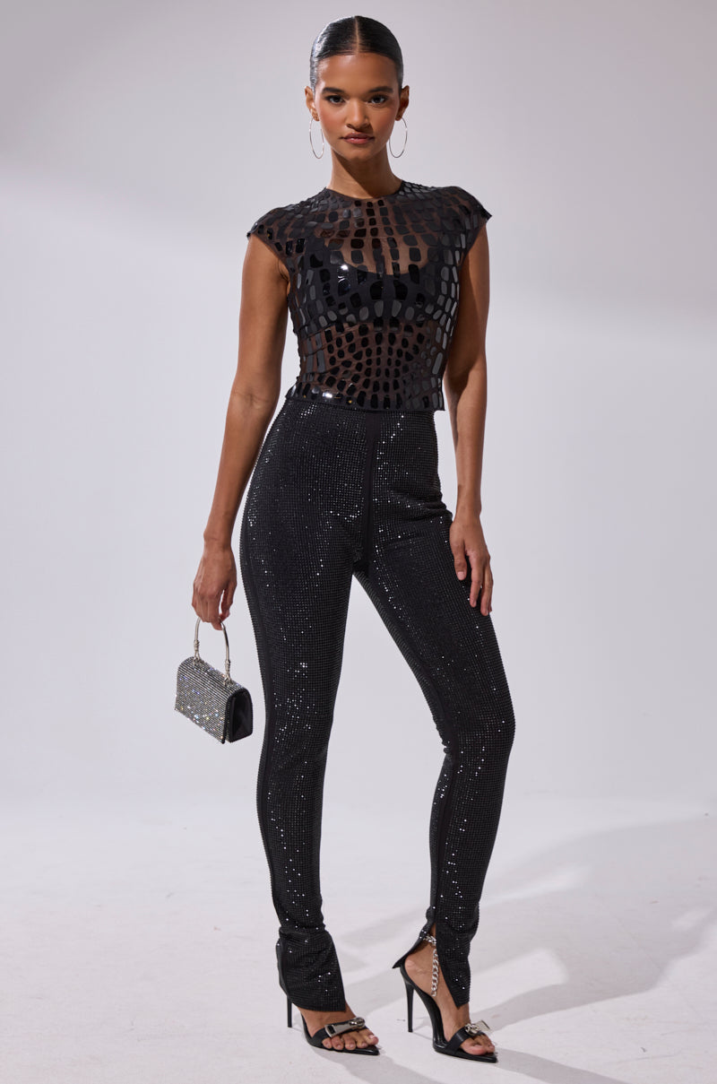 A woman poses against a plain background wearing the SPARKLE QUEEN RHINESTONE LEGGING, a black sheer patterned top, strappy black heels, and holding a small silver handbag.
