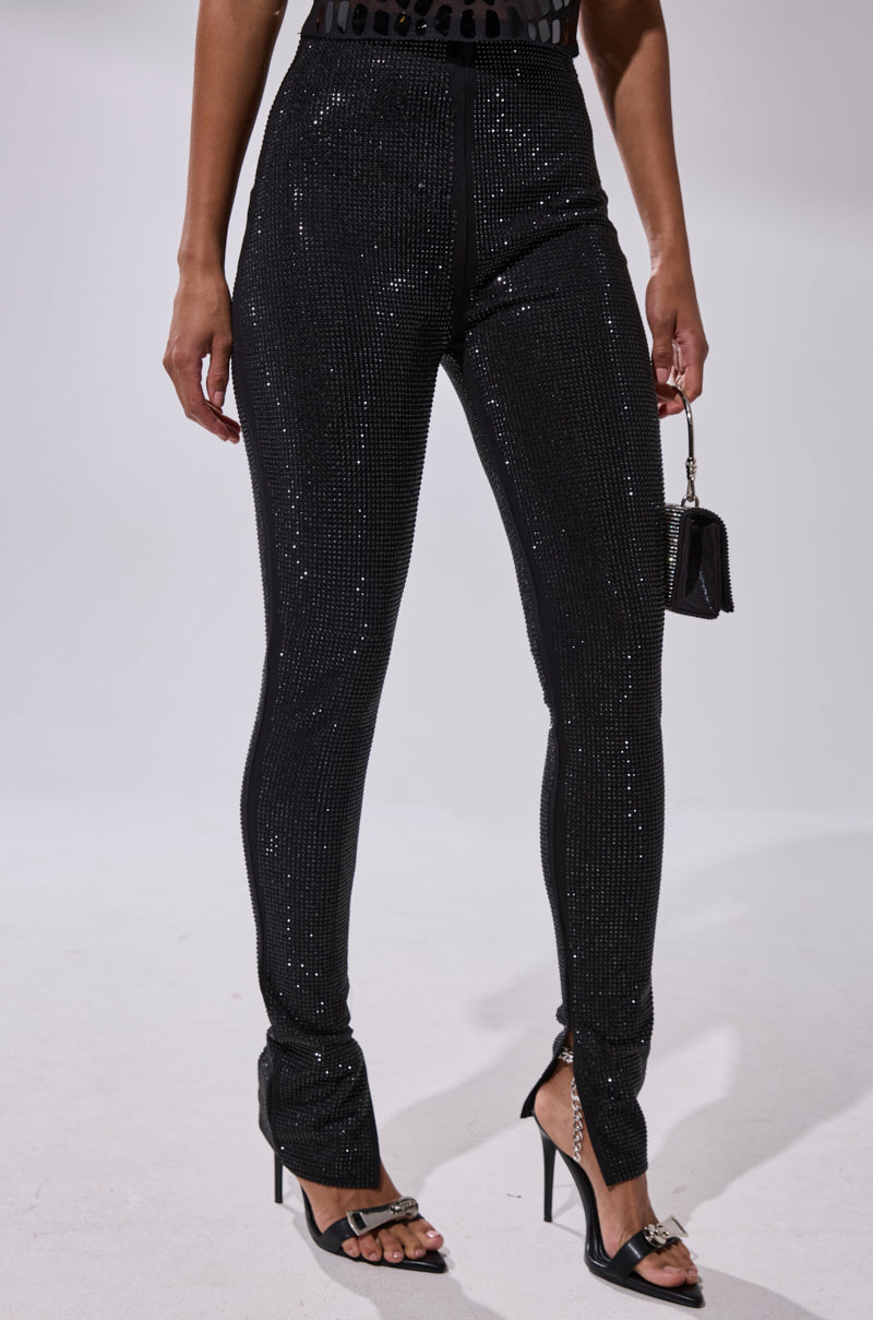 A person wears SPARKLE QUEEN RHINESTONE LEGGING with black heeled sandals and holds a small black handbag. The plain, light background and shoulder-down crop emphasize the leggings' sparkling design.
