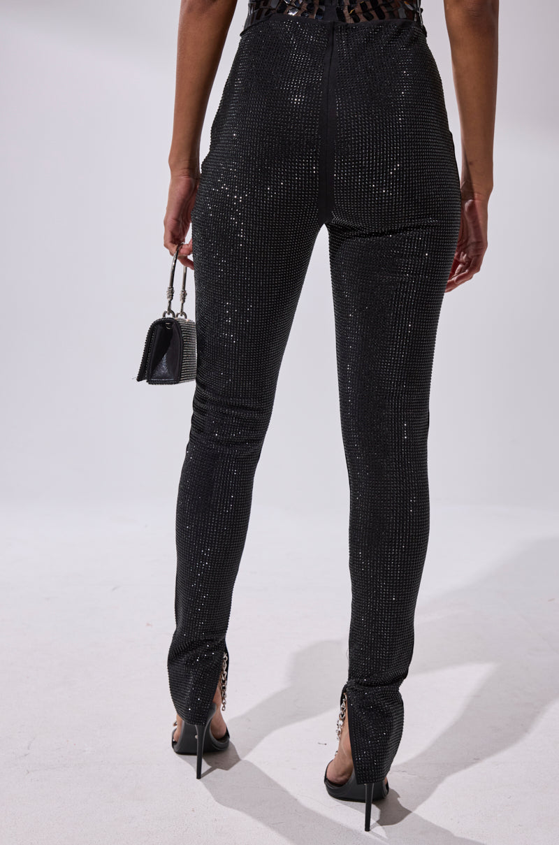 A person faces away from the camera, wearing SPARKLE QUEEN RHINESTONE LEGGING that shimmers with every move, paired with a small black handbag and sleek black heels.