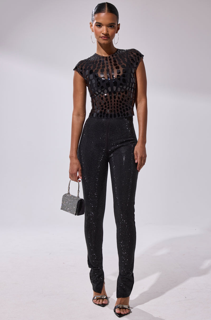 A woman poses against a plain backdrop in a black sheer top with circular patterns, the SPARKLE QUEEN RHINESTONE LEGGING, black heeled sandals, hoop earrings, and a small silver handbag.