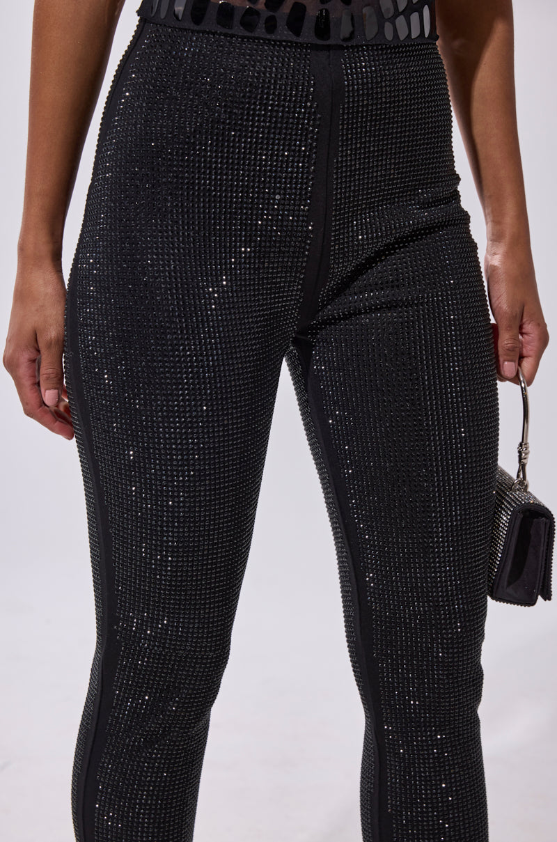 A person wears the SPARKLE QUEEN RHINESTONE LEGGING—black, high-waisted, and embellished with rhinestones—while holding a small metallic handbag. The image highlights the fitted pants from the waist to the legs.