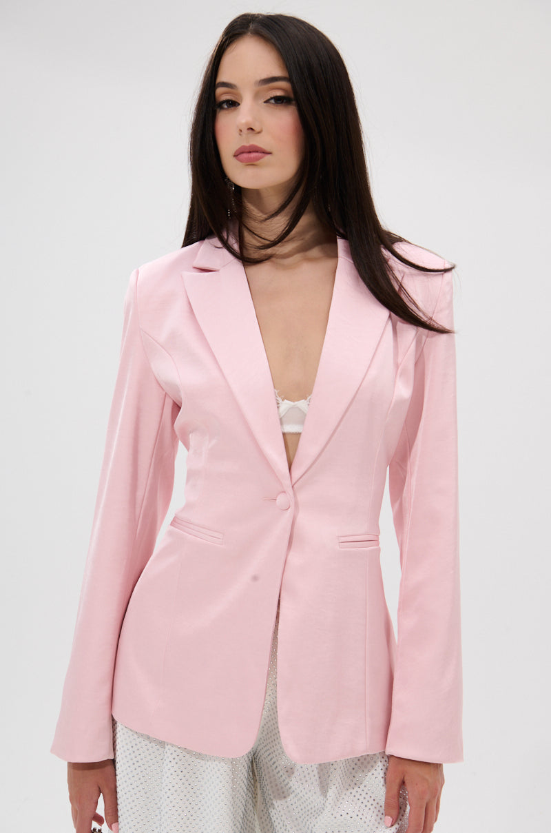 A woman with long dark hair models the DON'T MAKE ME BLUSH SATIN BLAZER in light pink over a white lace bra and white pants, standing against a plain light background. 1