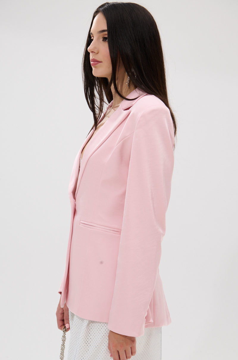 Woman with long dark hair wearing the DON’T MAKE ME BLUSH SATIN BLAZER in light pink, paired with a white skirt, standing in profile against a plain white background. 3