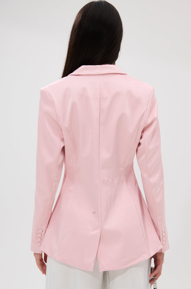 A woman with long dark hair is shown from the back wearing the DON'T MAKE ME BLUSH SATIN BLAZER—a light pink structured blazer with a rear vent and buttoned cuffs—paired with white pants against a plain, light-colored background. 4