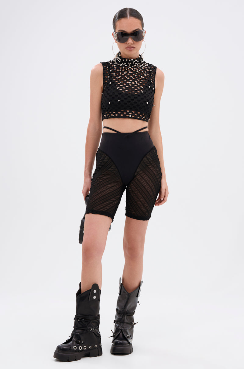 A woman poses against a white background wearing black sunglasses, a sequined crop top, strappy bikini bottoms, black combat boots with spikes, and the YAS QUEEN MESH BIKER SHORT—textured sheer mesh biker shorts with a high-waisted fit.