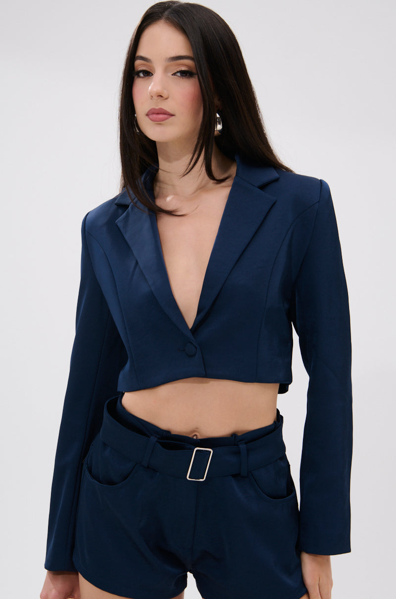 A woman with long dark hair wears the WILLA CROPPED BLAZER in navy blue with a deep V neckline and matching belted high-waisted shorts, highlighting a tailored look against a plain light background. 1