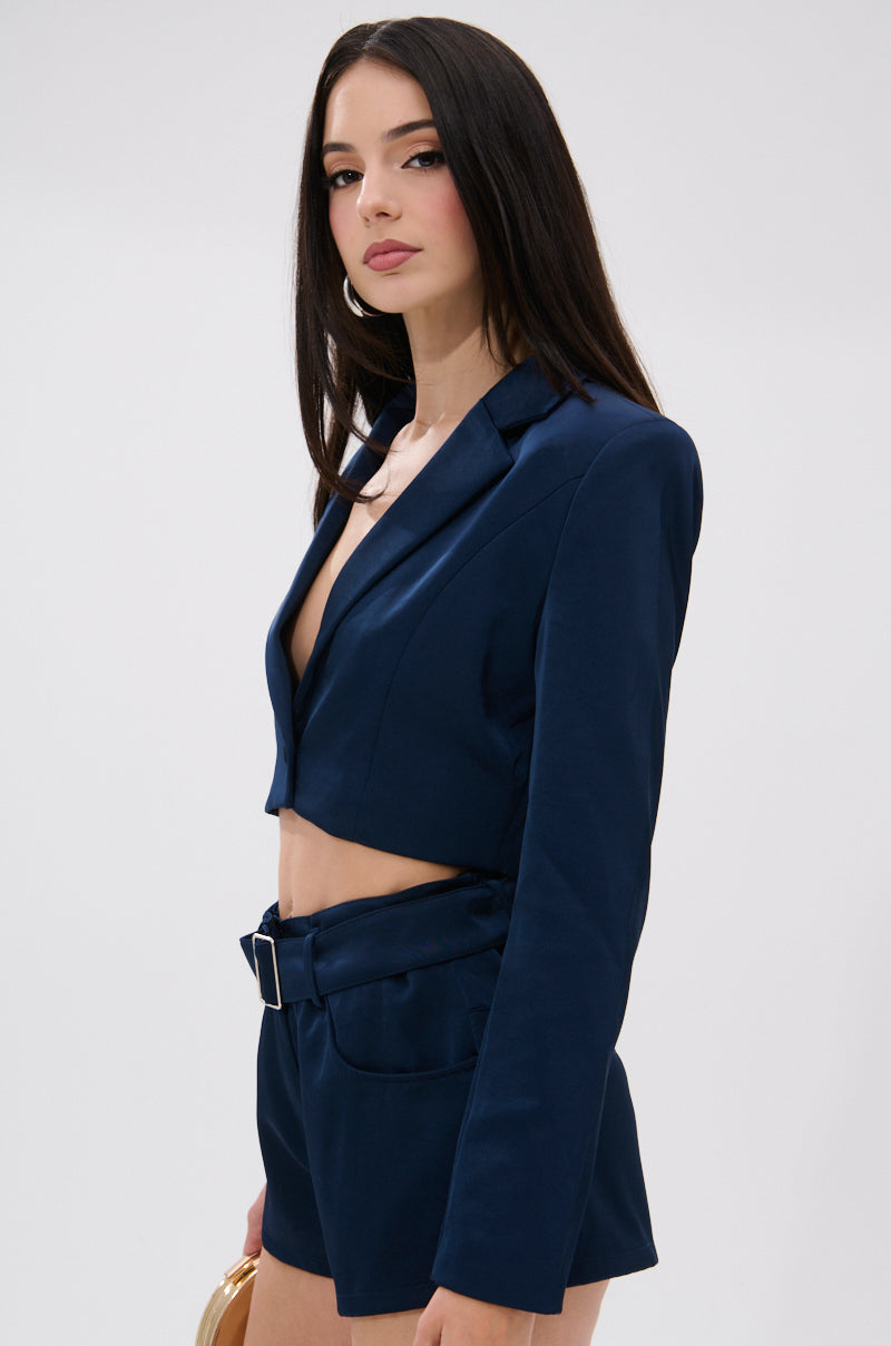 A woman with long straight hair wears the WILLA CROPPED BLAZER and matching high-waisted shorts with a belt, holding a tan clutch and looking confidently at the camera against a plain light background. 2