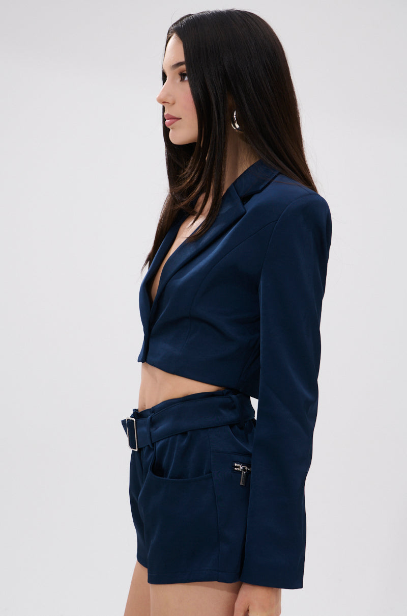 A woman with long dark hair wears the WILLA CROPPED BLAZER and matching high-waisted belted shorts, highlighting a sleek tailored silhouette as she stands in profile against a plain light background. 3
