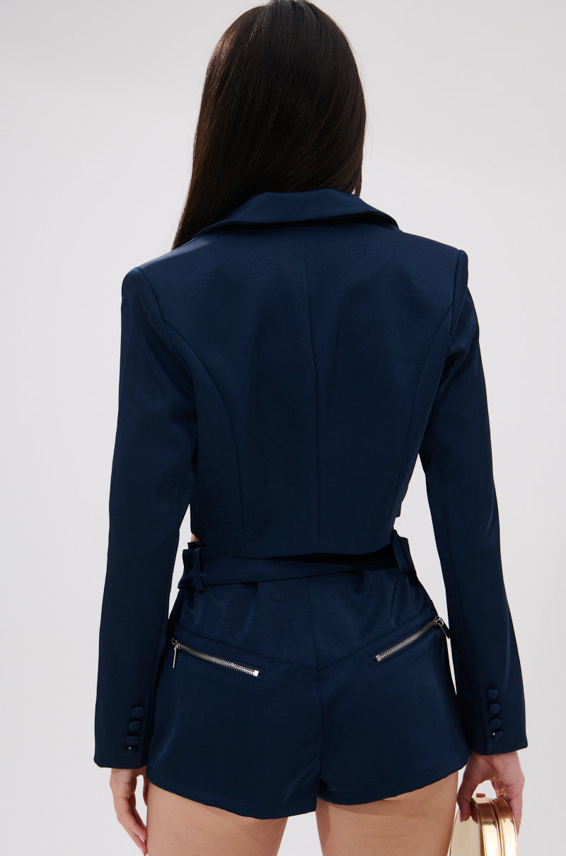 A woman with long dark hair, seen from behind, wears the WILLA CROPPED BLAZER in navy blue with matching high-waisted shorts featuring zipper details, and carries a round gold handbag in her left hand. 4