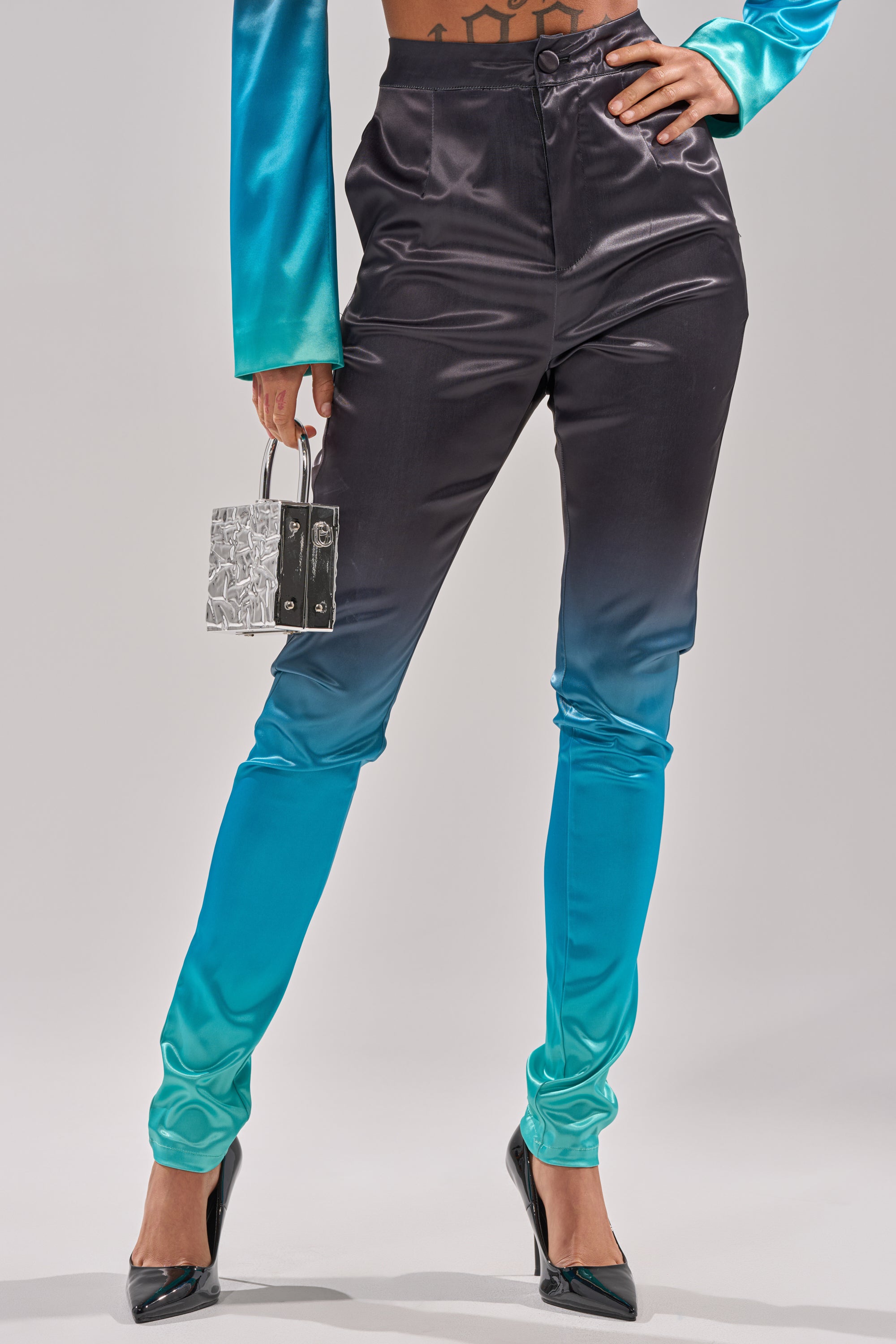 A person wears the NIGHT AND DAY OMBRE SKINNY TROUSER, featuring a high-rise fit and ombre fade from black to teal, paired with a matching top, black heels, and a silver handbag; photo cropped at the shoulders. 1