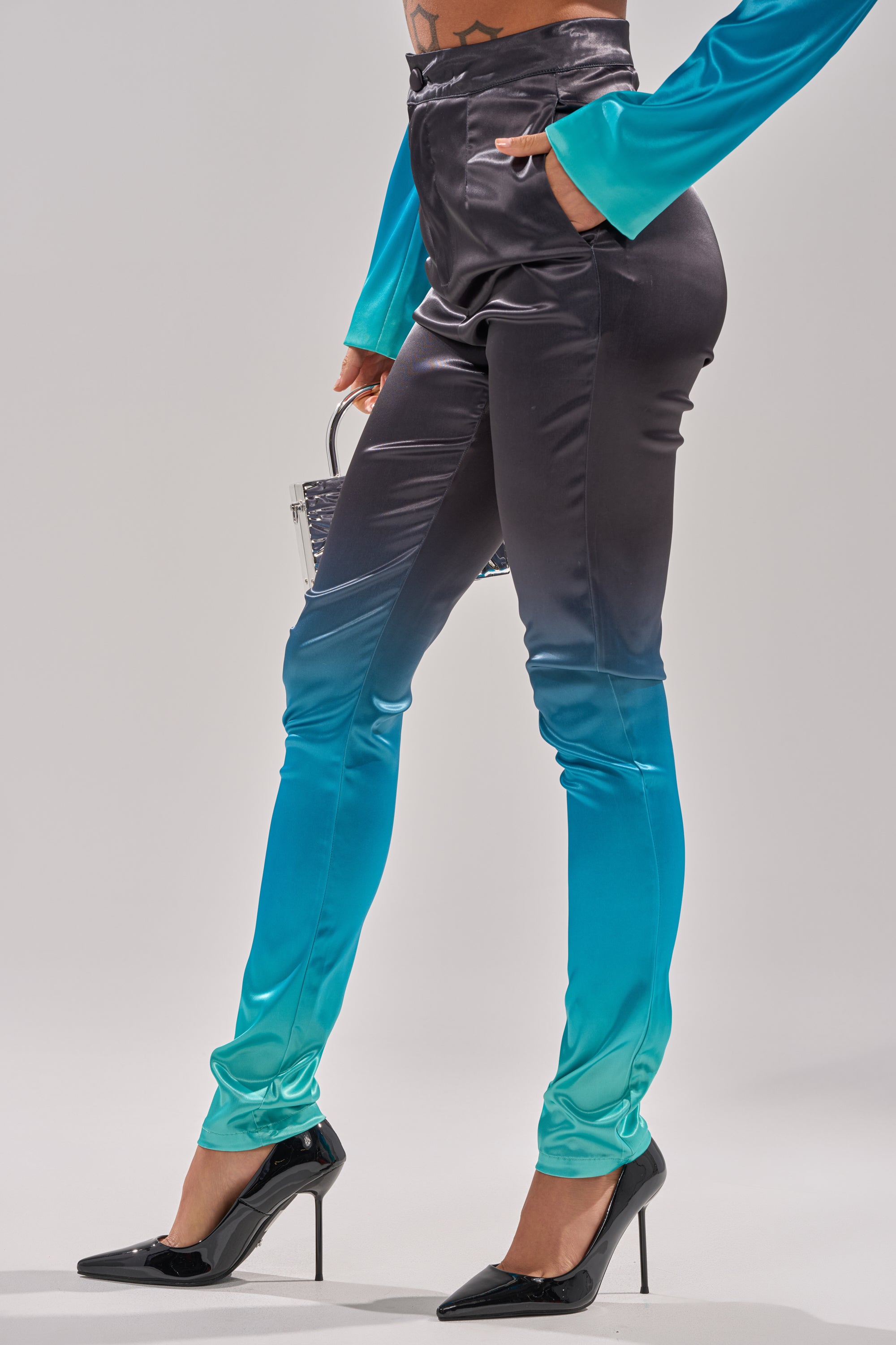 Wearing the NIGHT AND DAY OMBRE SKINNY TROUSER with a shiny black-to-turquoise ombre effect, matched with a turquoise top, black high heels, and a small bag against a plain background. 3