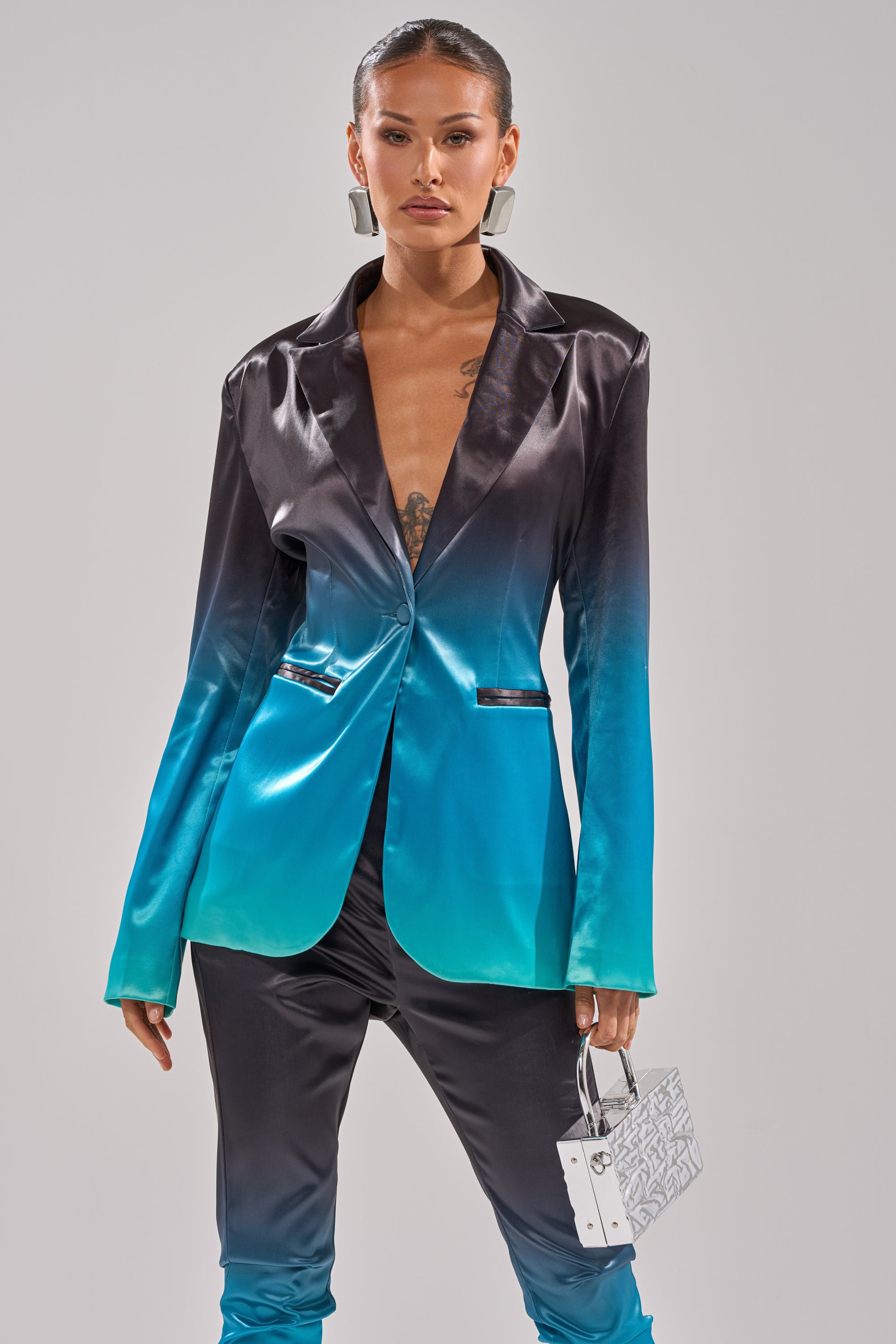 Wearing the NIGHT AND DAY OMBRE BLAZER and matching pants, which fade from black to teal, they style the look with a small white handbag and bold silver earrings, posing against a plain background. 0