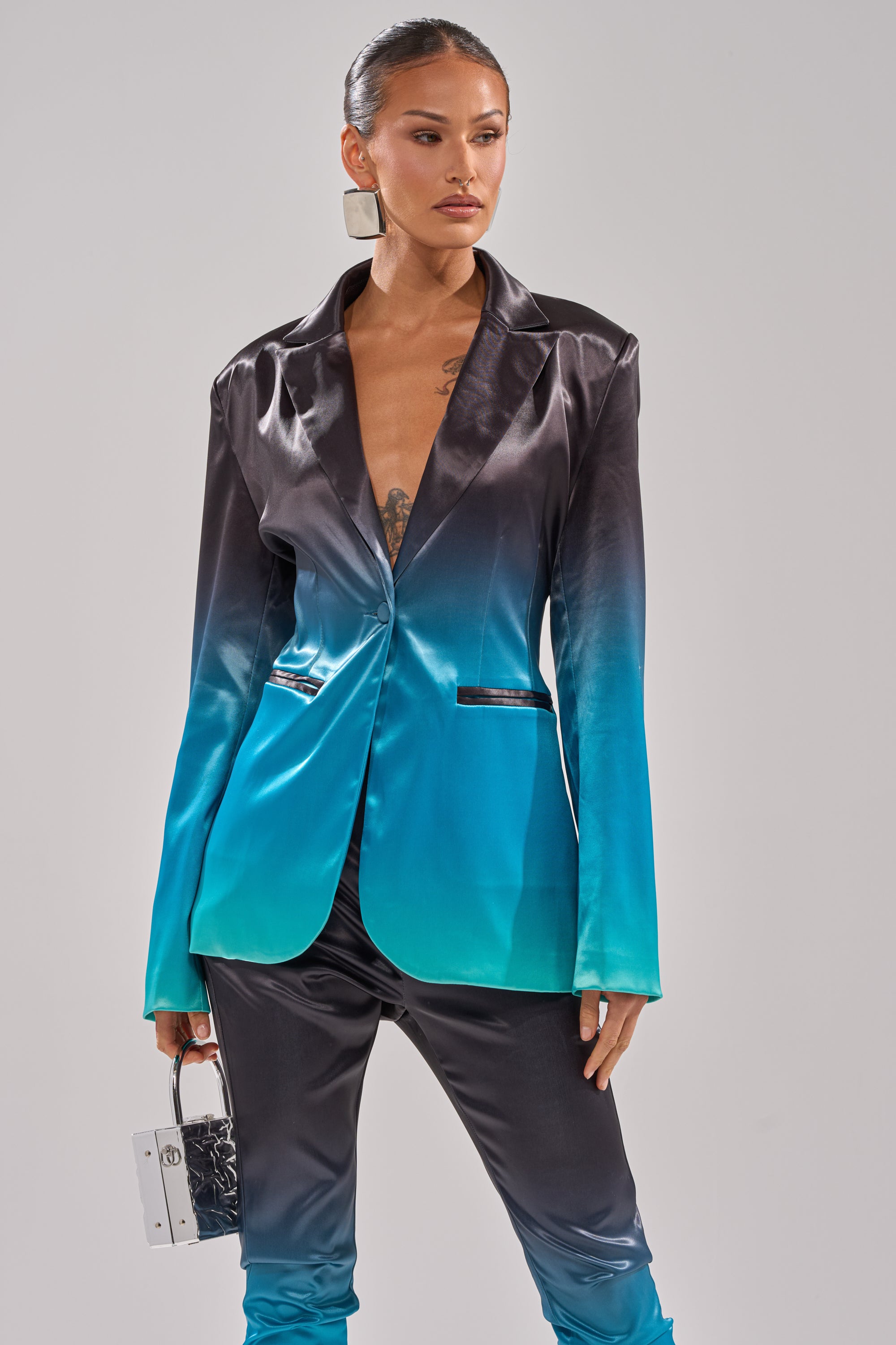 Wearing the NIGHT AND DAY OMBRE BLAZER with matching longline pants in a black-to-turquoise gradient, a woman holds a small purse and sports pulled-back hair, radiating confidence and modern style. 2