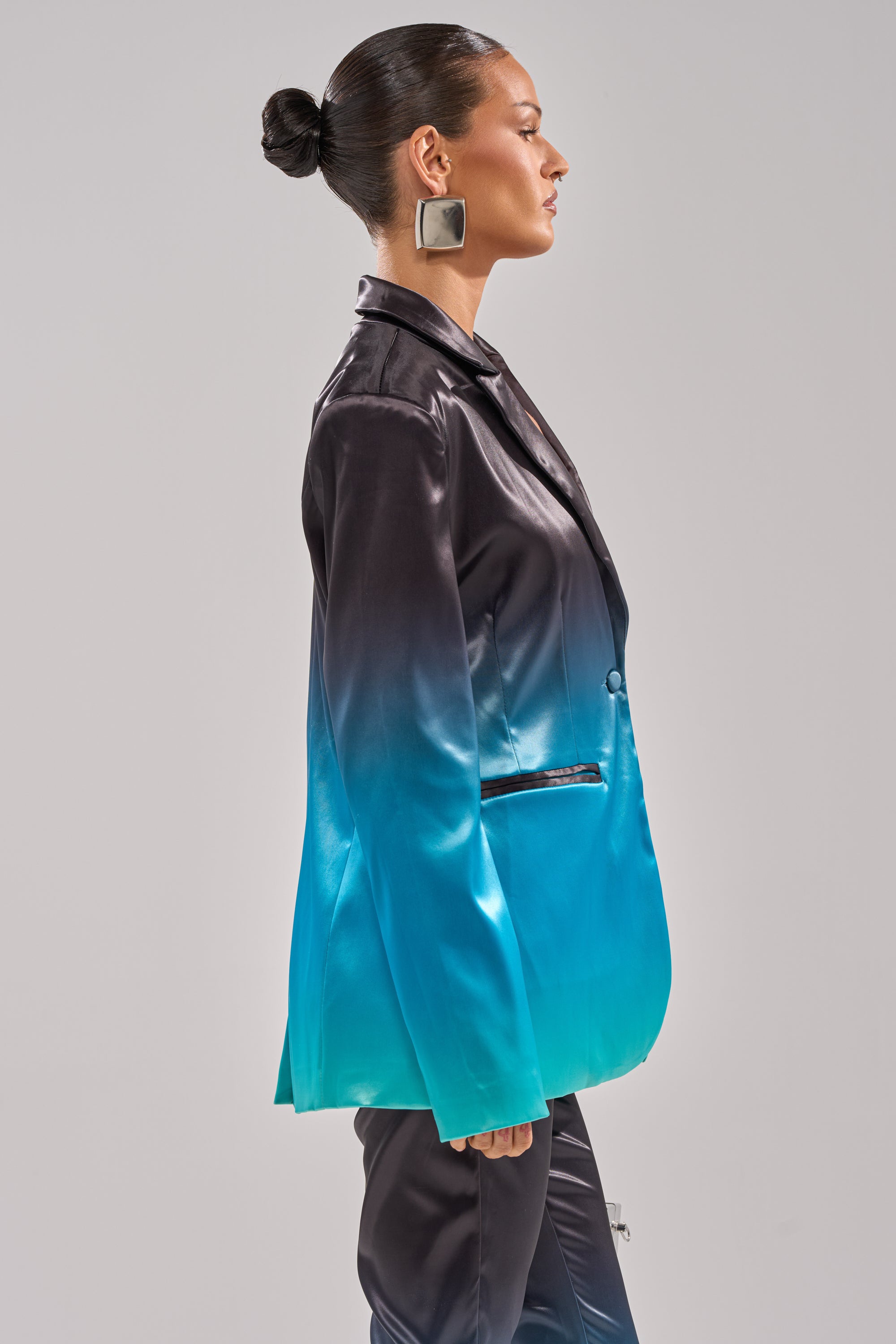 A woman with dark hair in a bun models the NIGHT AND DAY OMBRE BLAZER, featuring a longline silhouette that fades from black at the shoulders to turquoise at the hem. She is shown in profile against a plain light background, wearing bold square earrings. 3