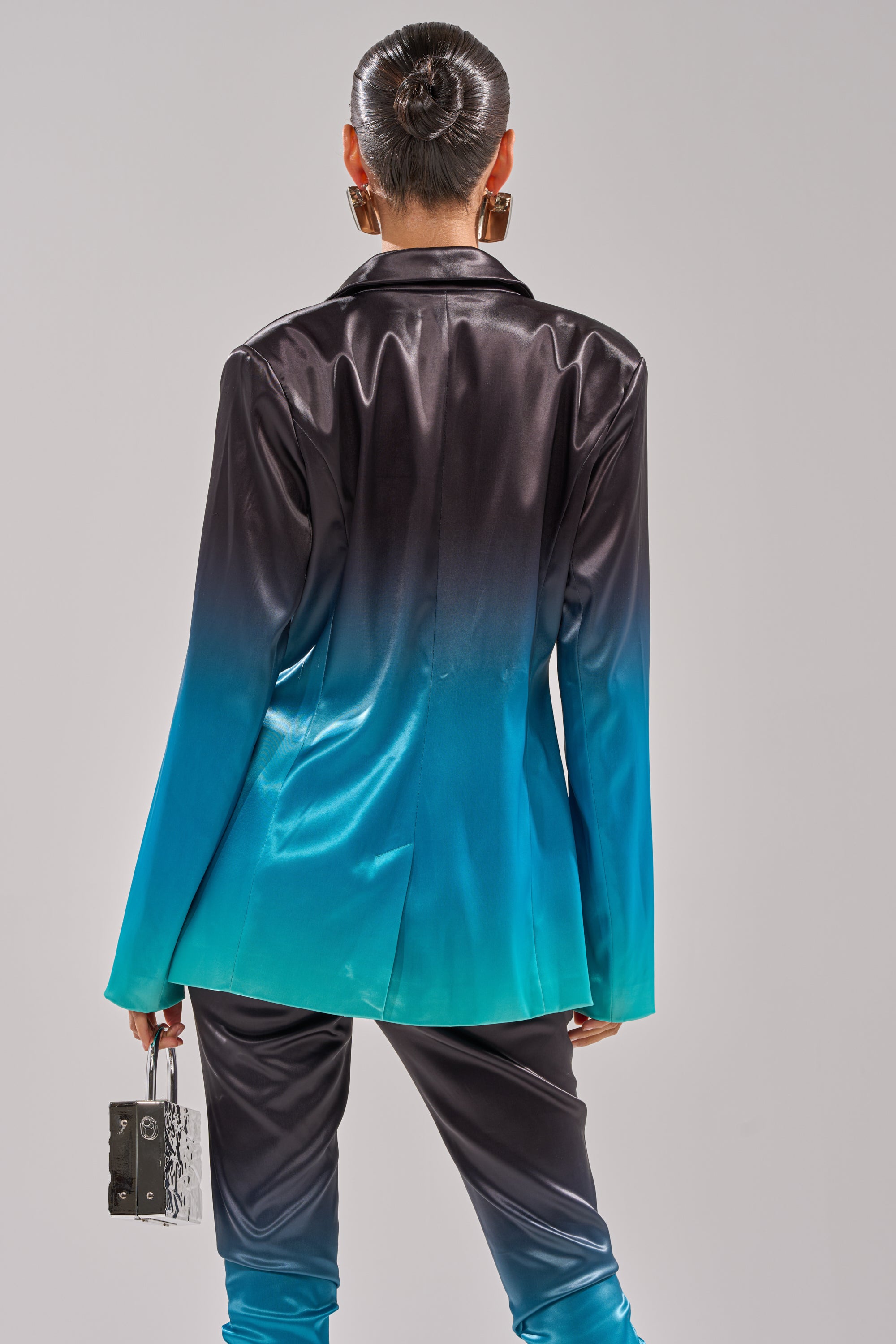 A person with a sleek bun wears the NIGHT AND DAY OMBRE BLAZER and matching pants that fade from black to bright teal. Holding a small rectangular purse, they stand facing away from the camera. 4
