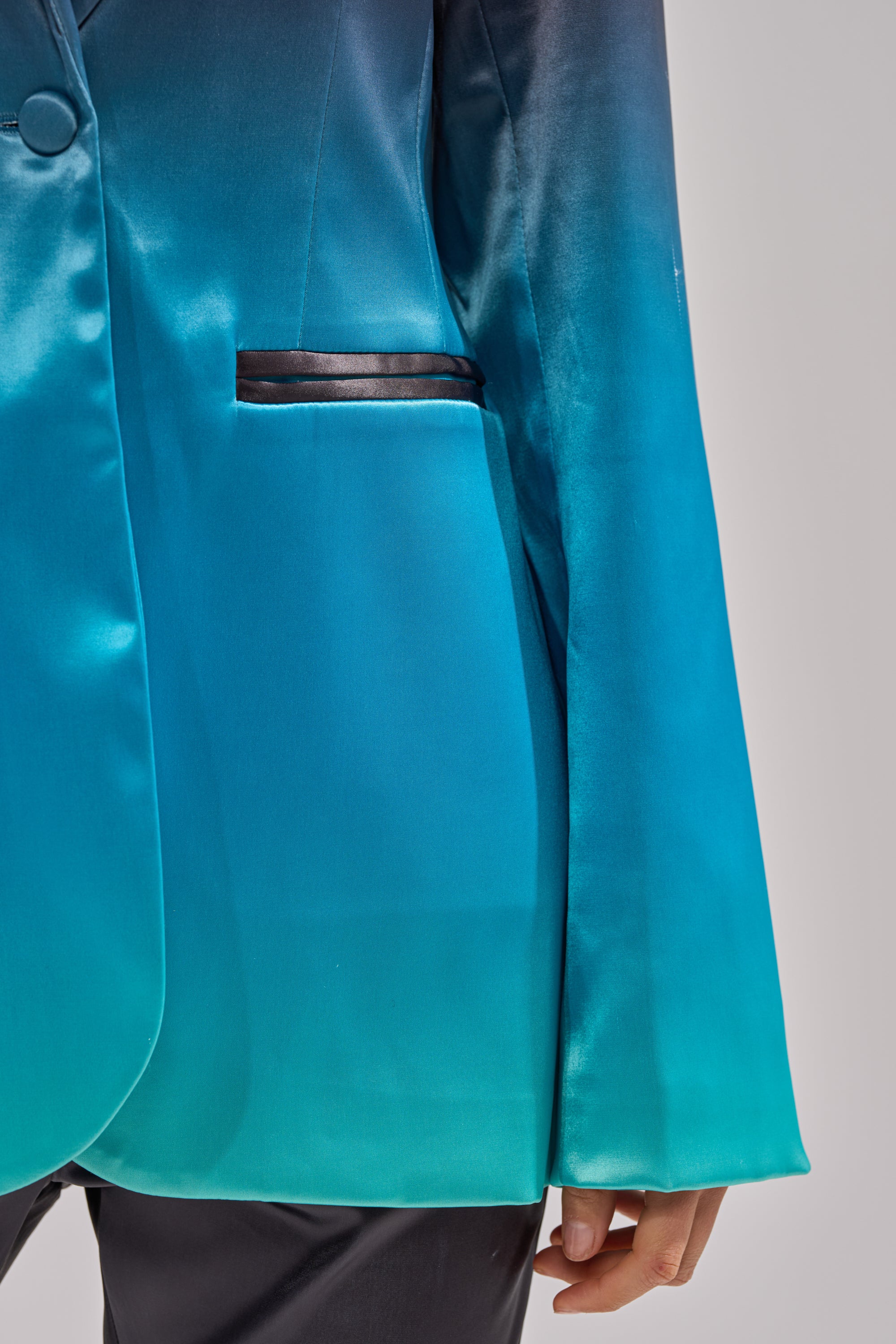 A close-up of a person wearing the NIGHT AND DAY OMBRE BLAZER in shiny teal and blue satin with black pocket trim, paired with black pants. Only the lower half of the blazer and the person’s hand are visible. 7