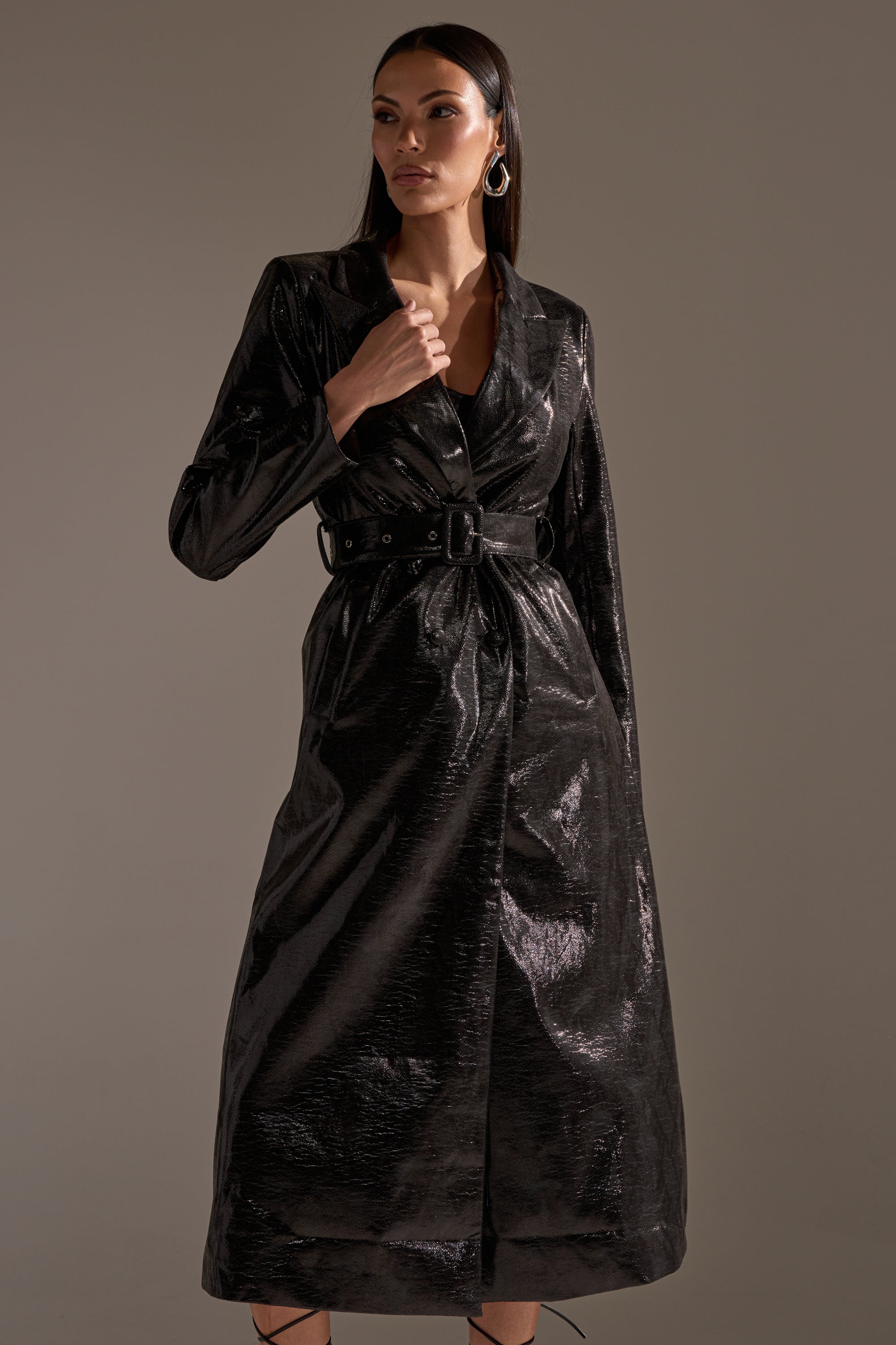 A woman with long dark hair and gold hoop earrings poses against a neutral background, holding the collar of the shiny black ACQUIRED TASTE FAUX LEATHER TRENCH by AKIRA Label.