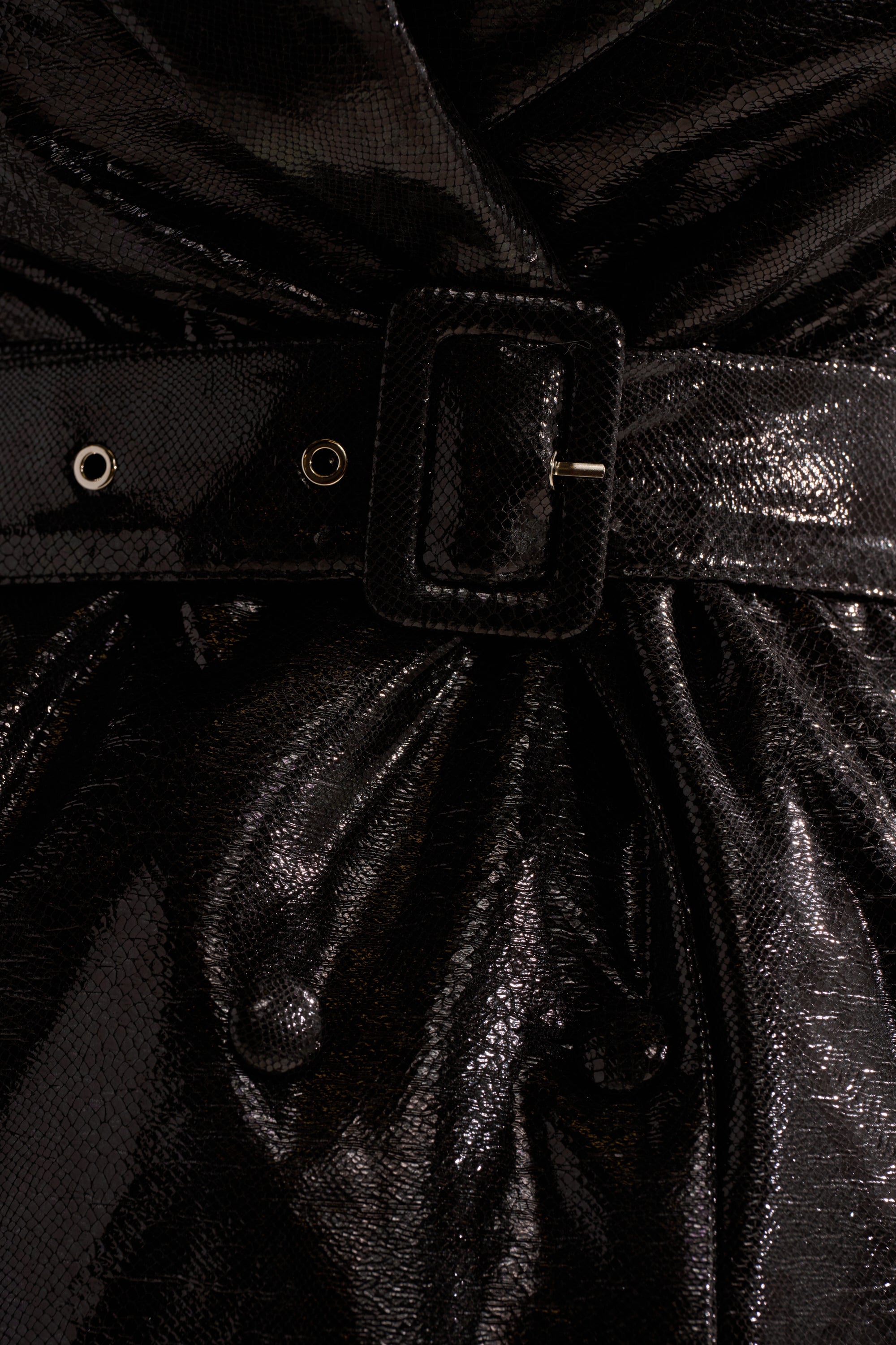 Close-up of the ACQUIRED TASTE FAUX LEATHER TRENCH in shiny black textured faux leather, cinched at the waist with a matching belt featuring a rectangular buckle and metal eyelets.