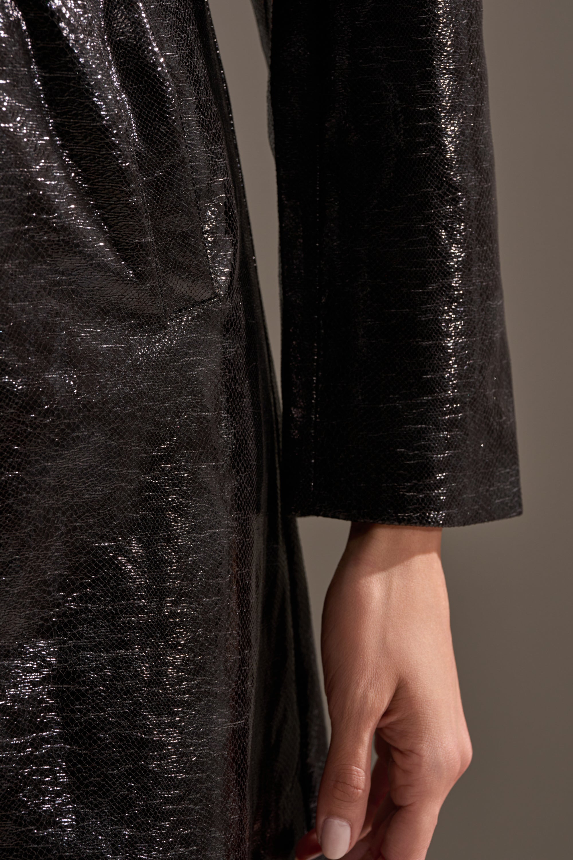 A close-up of a hand resting by the side, wearing the black, shiny ACQUIRED TASTE FAUX LEATHER TRENCH with long sleeves. The background is plain and neutral.
