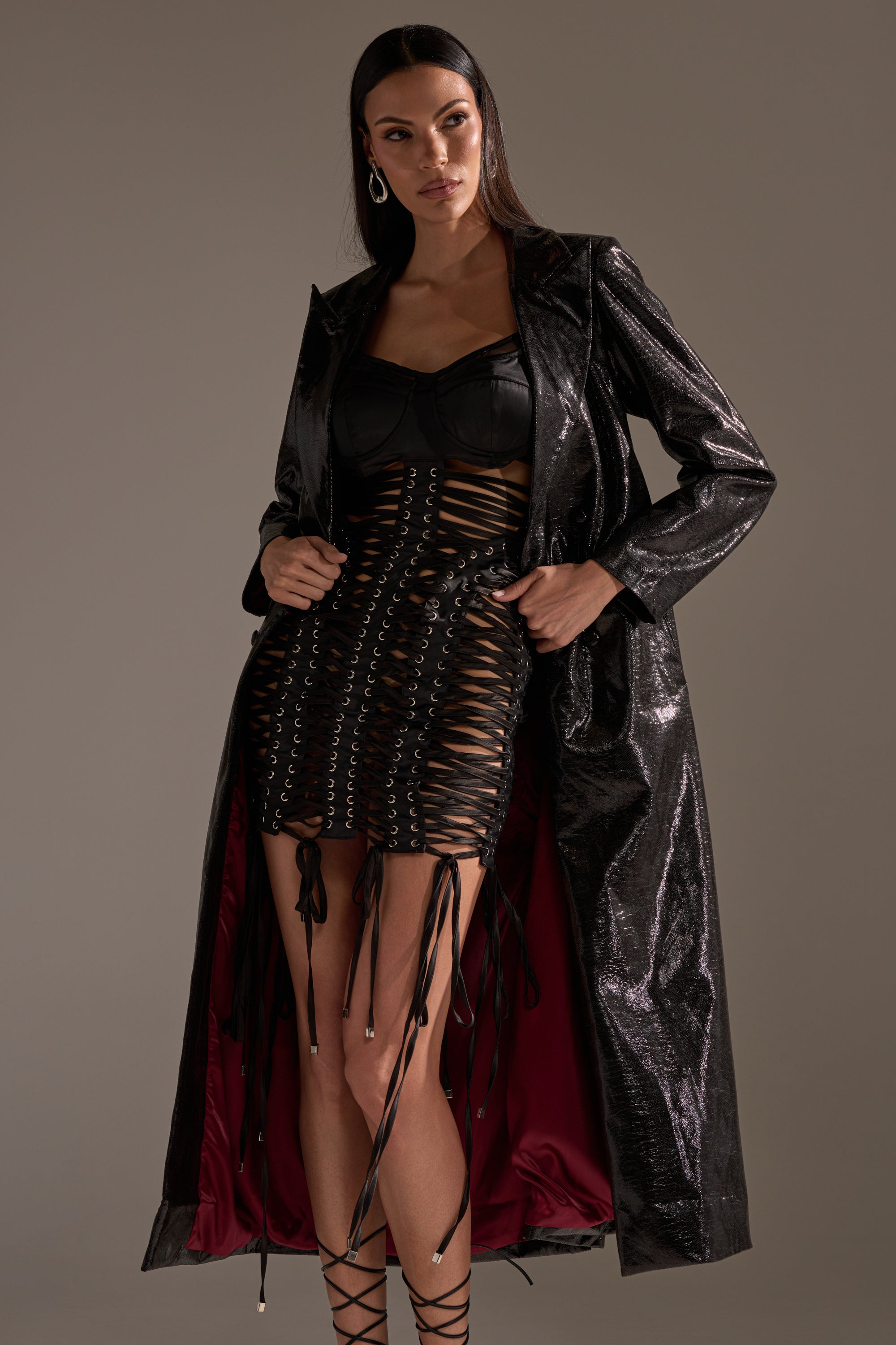 A woman stands confidently in the shiny black ACQUIRED TASTE FAUX LEATHER TRENCH with dark red lining, paired with a black bustier and crisscross lace-up mini skirt. Her long dark hair and hoop earrings finish the bold look.