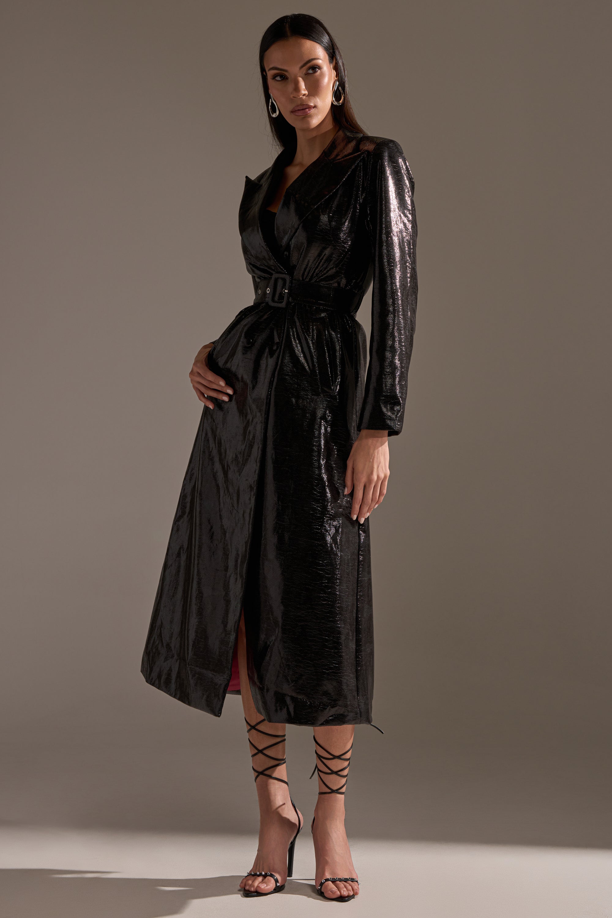 A woman exudes confidence in the ACQUIRED TASTE FAUX LEATHER TRENCH, a shiny black belted midi coat, styled with strappy high-heeled sandals and large hoop earrings against a neutral backdrop.