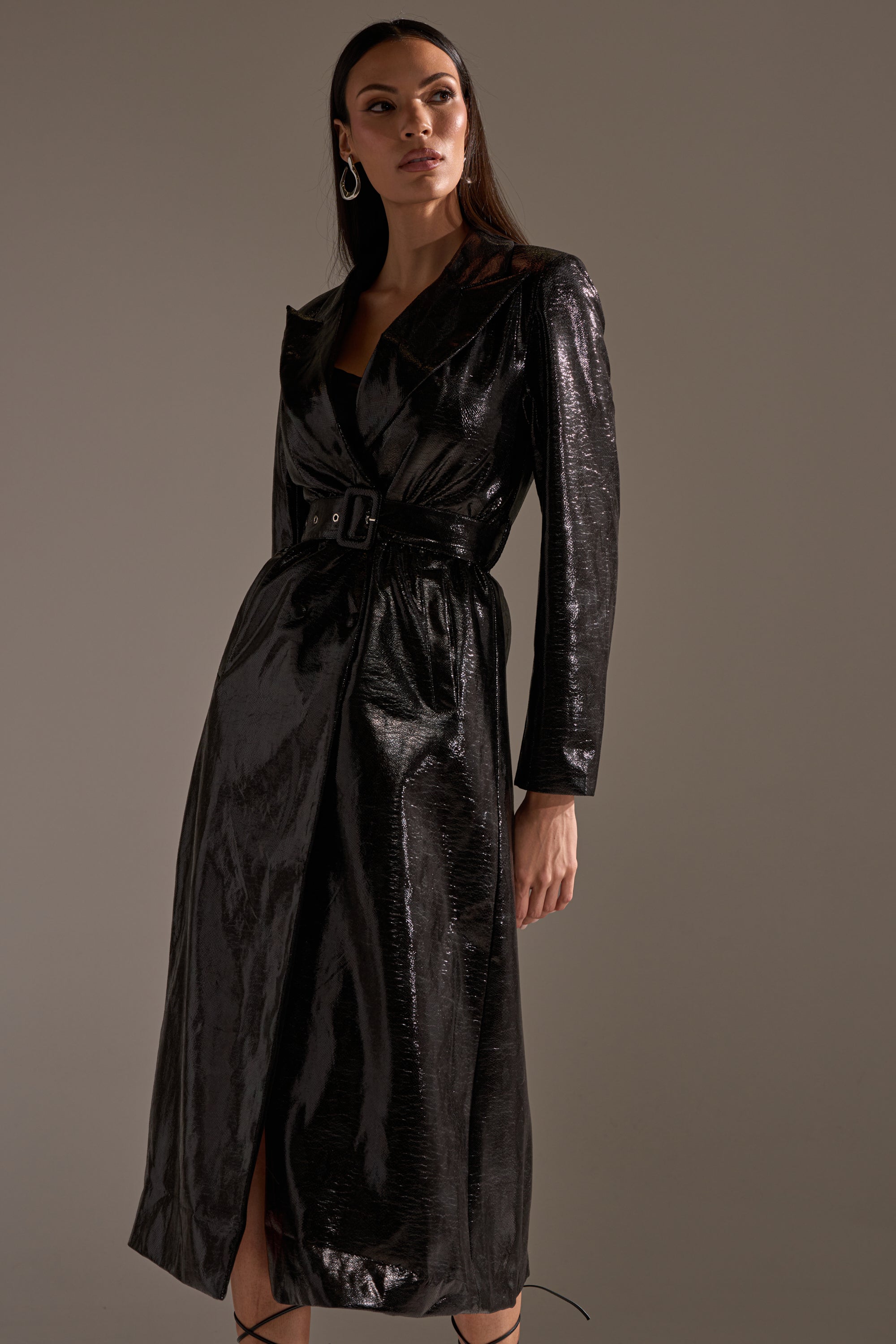 A woman models the ACQUIRED TASTE FAUX LEATHER TRENCH with long sleeves and a sleek belt, posing against a plain grey background.
