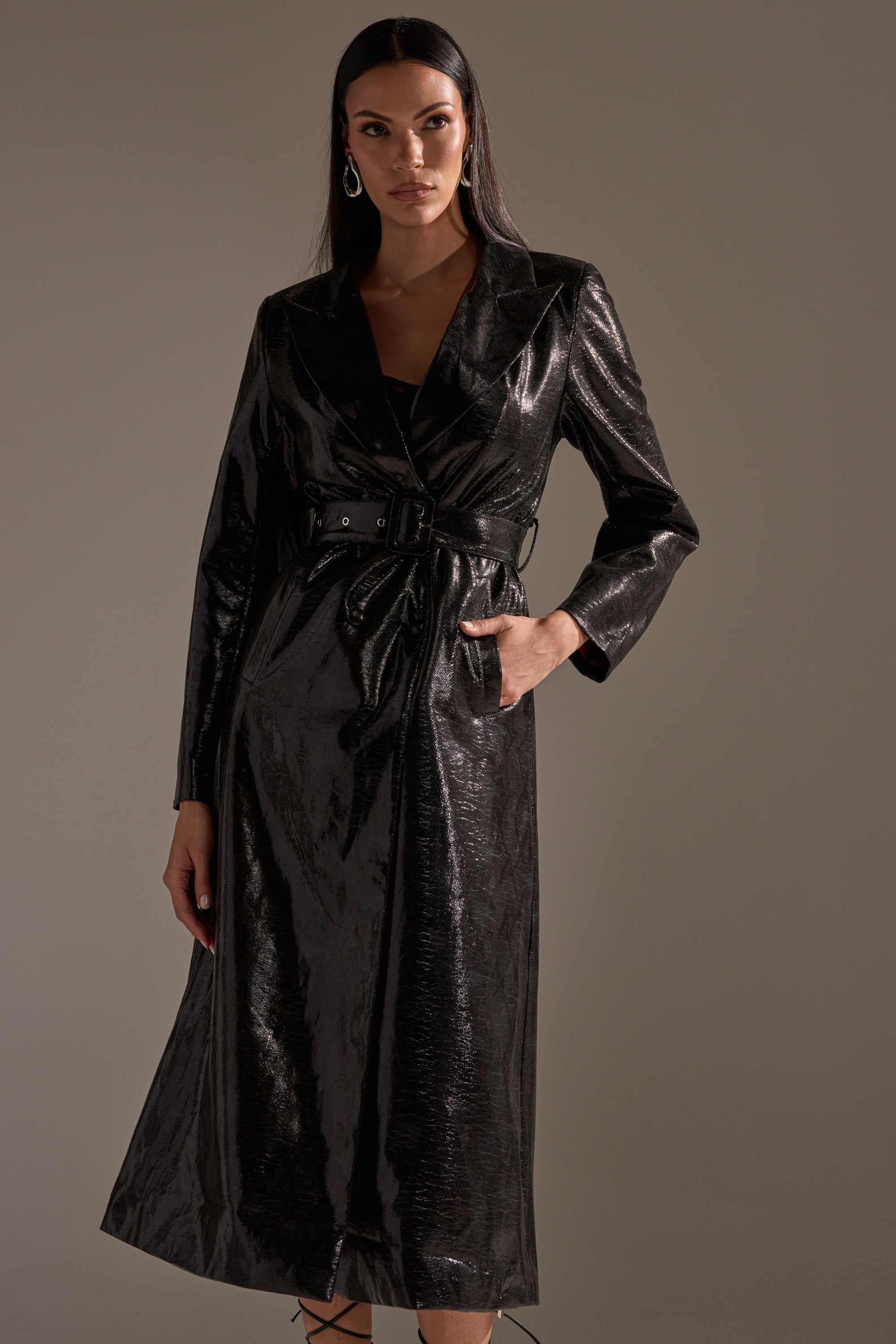 A woman models the ACQUIRED TASTE FAUX LEATHER TRENCH by AKIRA Label, a longline coat with a shiny, textured finish. She has straight dark hair, wears hoop earrings, and poses with one hand in her pocket against a plain background.