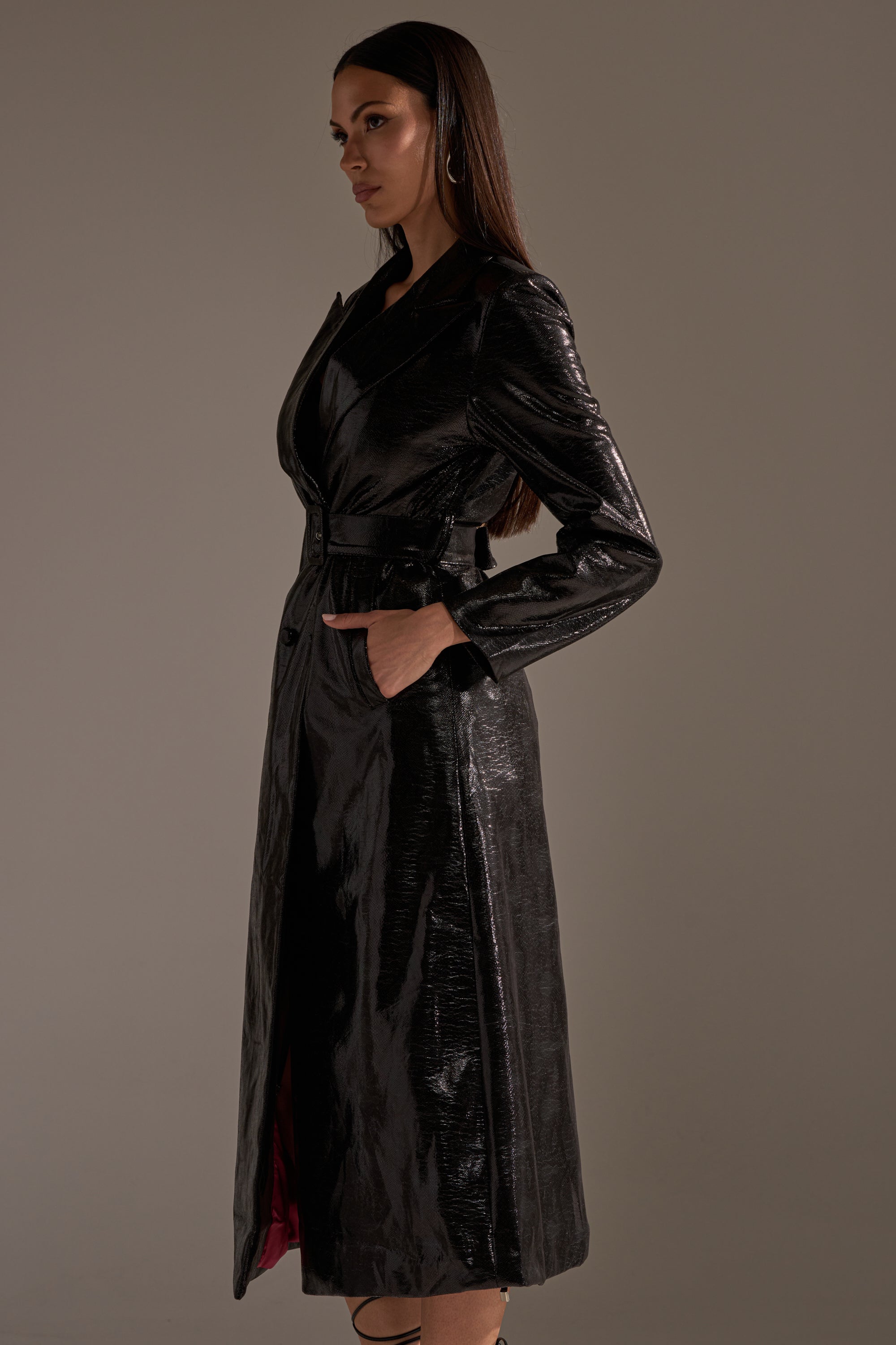 A woman stands in profile with her hands in the pockets of the ACQUIRED TASTE FAUX LEATHER TRENCH by AKIRA Label. The shiny black, belted coat contrasts with her long, straight dark hair and a plain, neutral background.