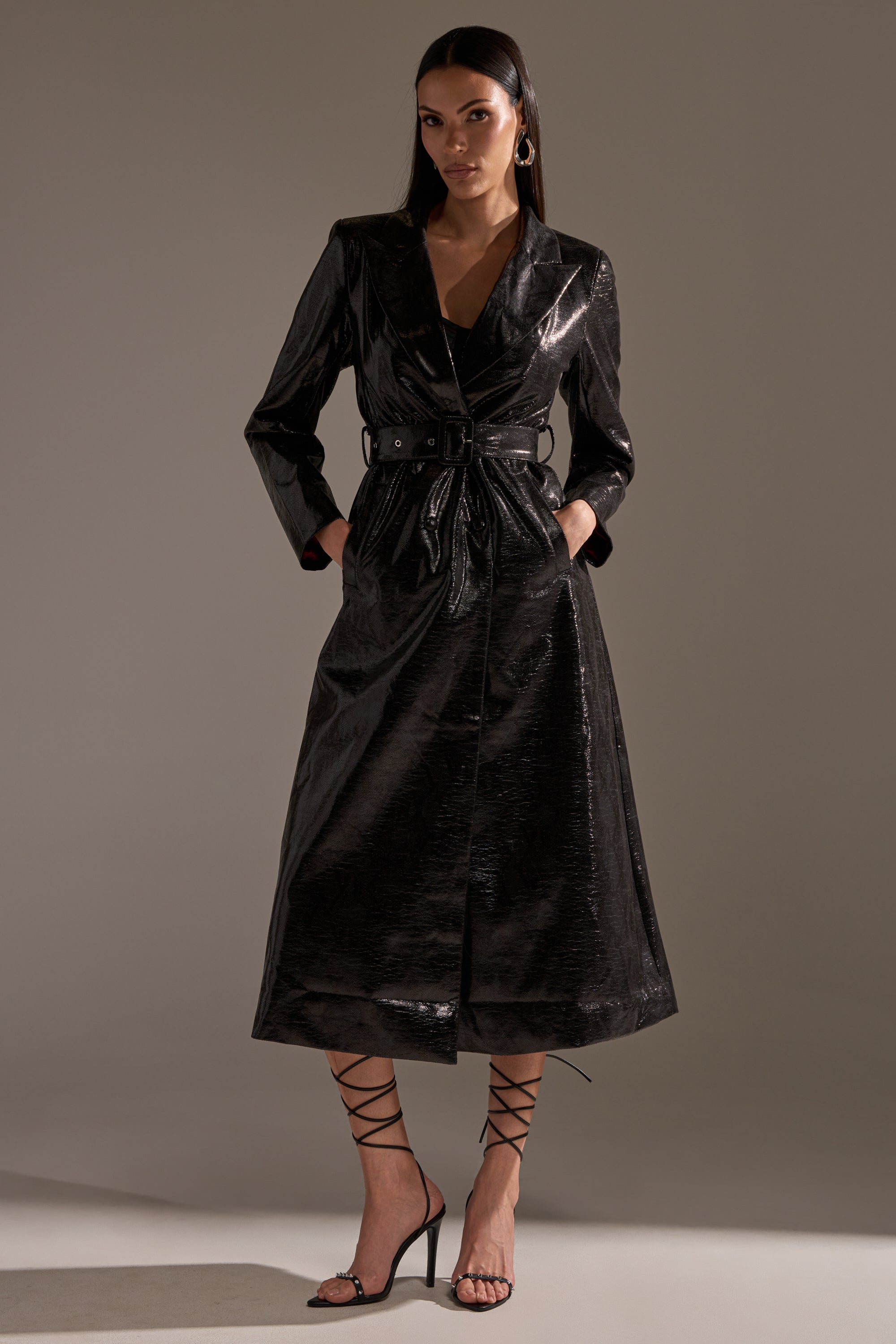 A woman stands confidently with hands in pockets, wearing the ACQUIRED TASTE FAUX LEATHER TRENCH featuring a glossy finish and belted waist, paired with strappy heels and hoop earrings against a neutral background.