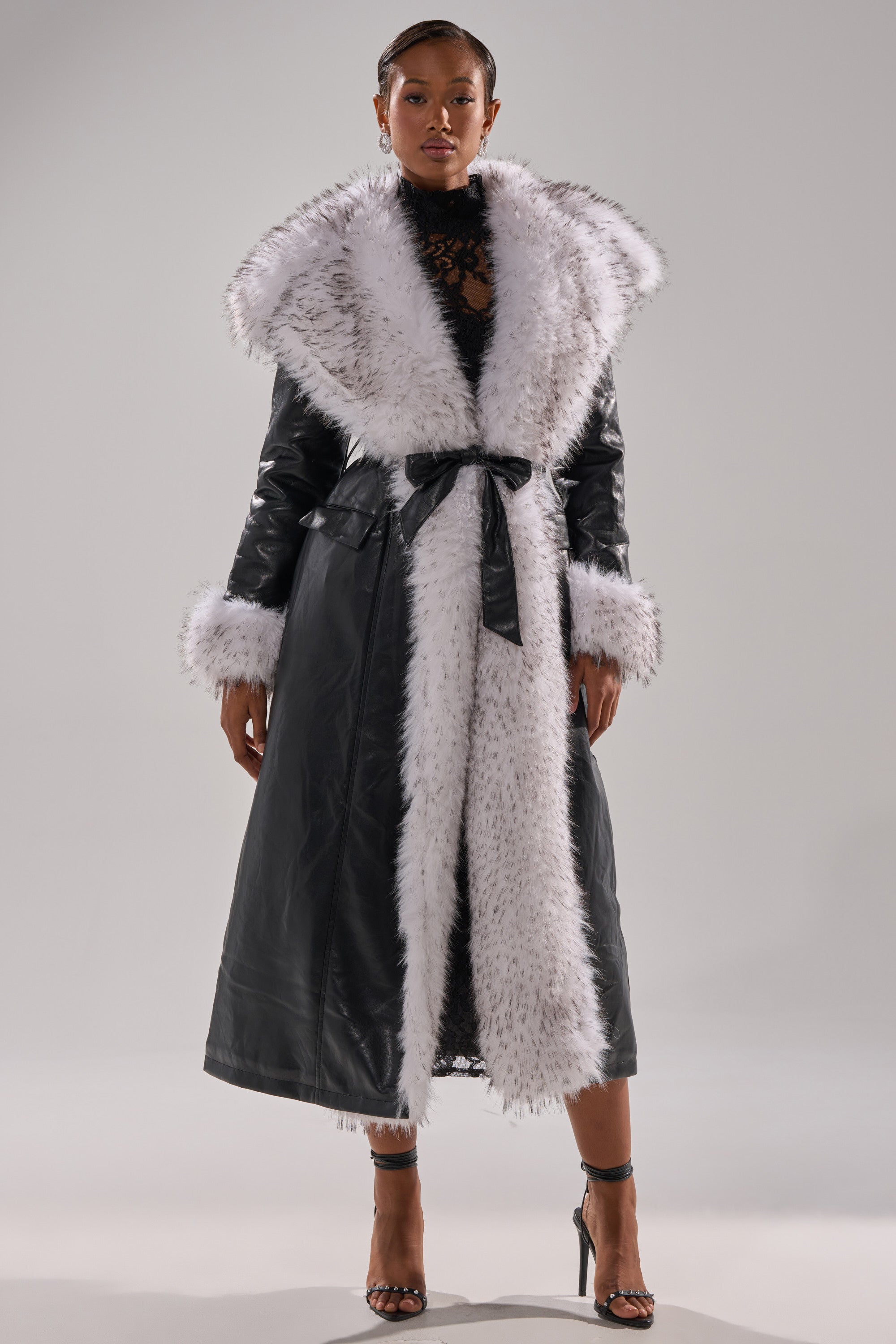 A woman exudes confidence in the FIONA BUTTER LUXE FUR TRIM TRENCH, featuring a wide white fur collar and cuffs, belted at the waist. She styles it with strappy heels and a black lace top for an eye-catching look.