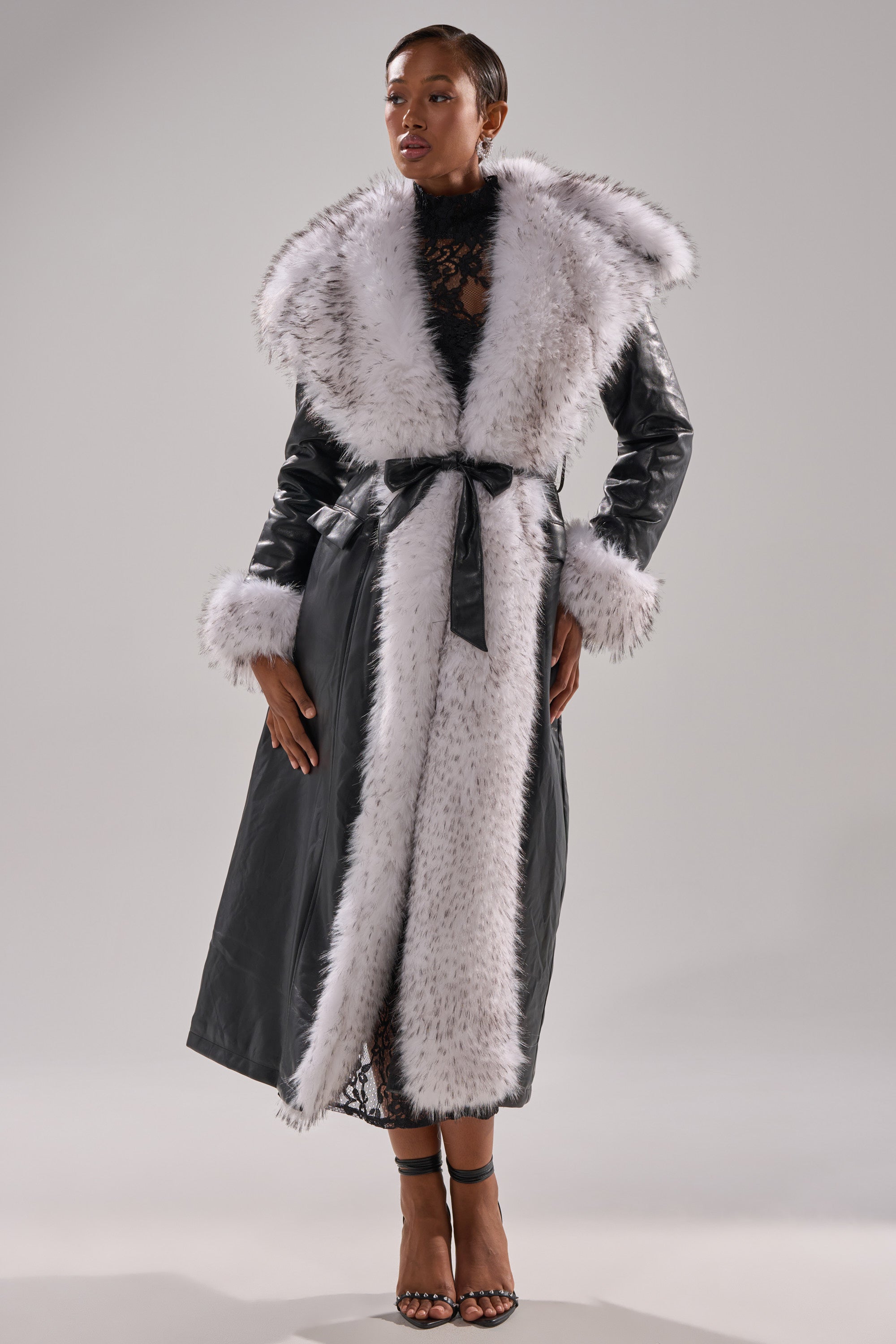 A woman models the FIONA BUTTER LUXE FUR TRIM TRENCH, featuring a longline silhouette with wide white fur collar, fur-trimmed cuffs, and a matching fur-lined front. She styles it with black heeled sandals and a black lace top underneath.