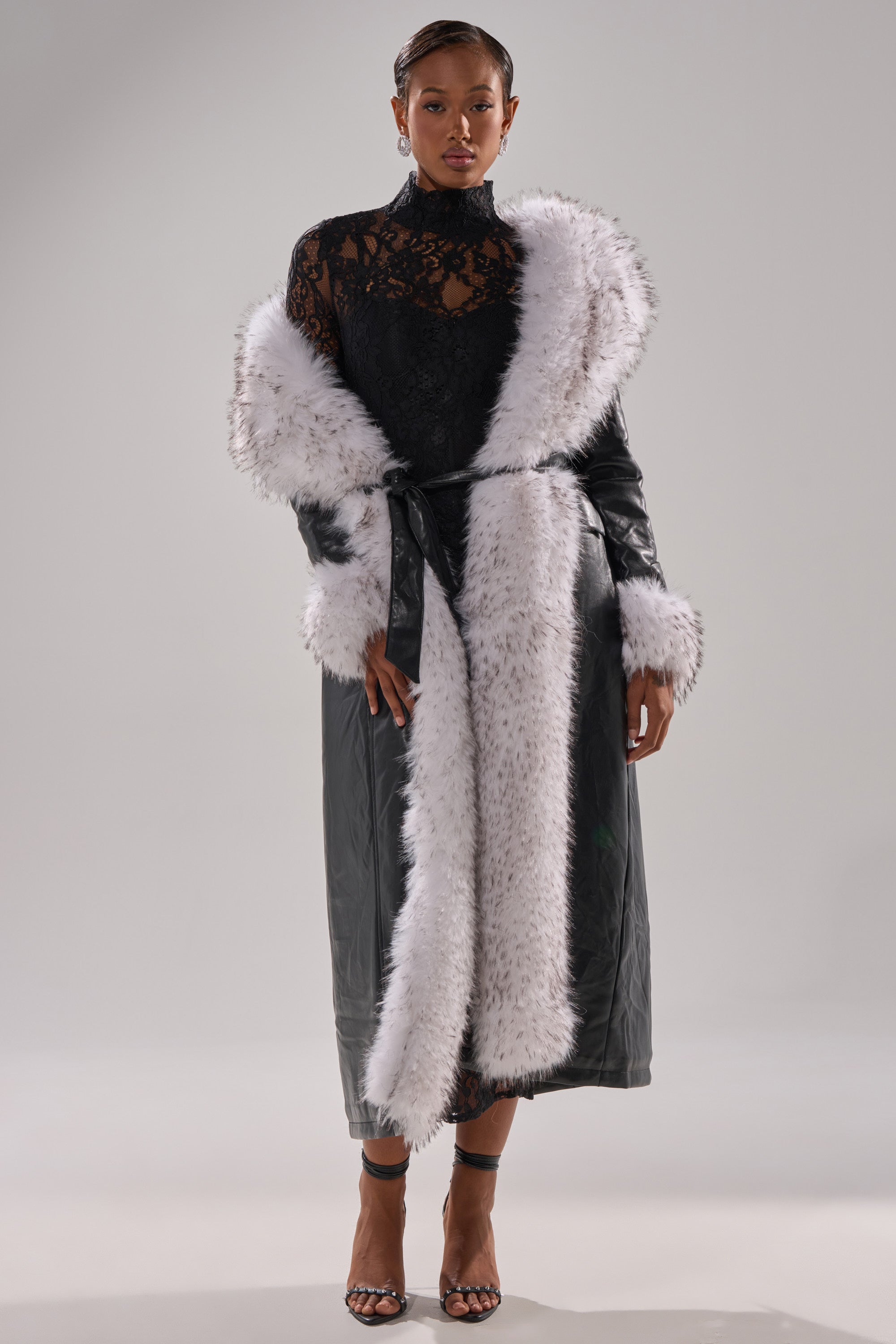 A woman poses confidently in the FIONA BUTTER LUXE FUR TRIM TRENCH, a longline faux leather coat with white fur trim at the collar, cuffs, and front. She pairs it with a black lace top and black heels against a plain background.
