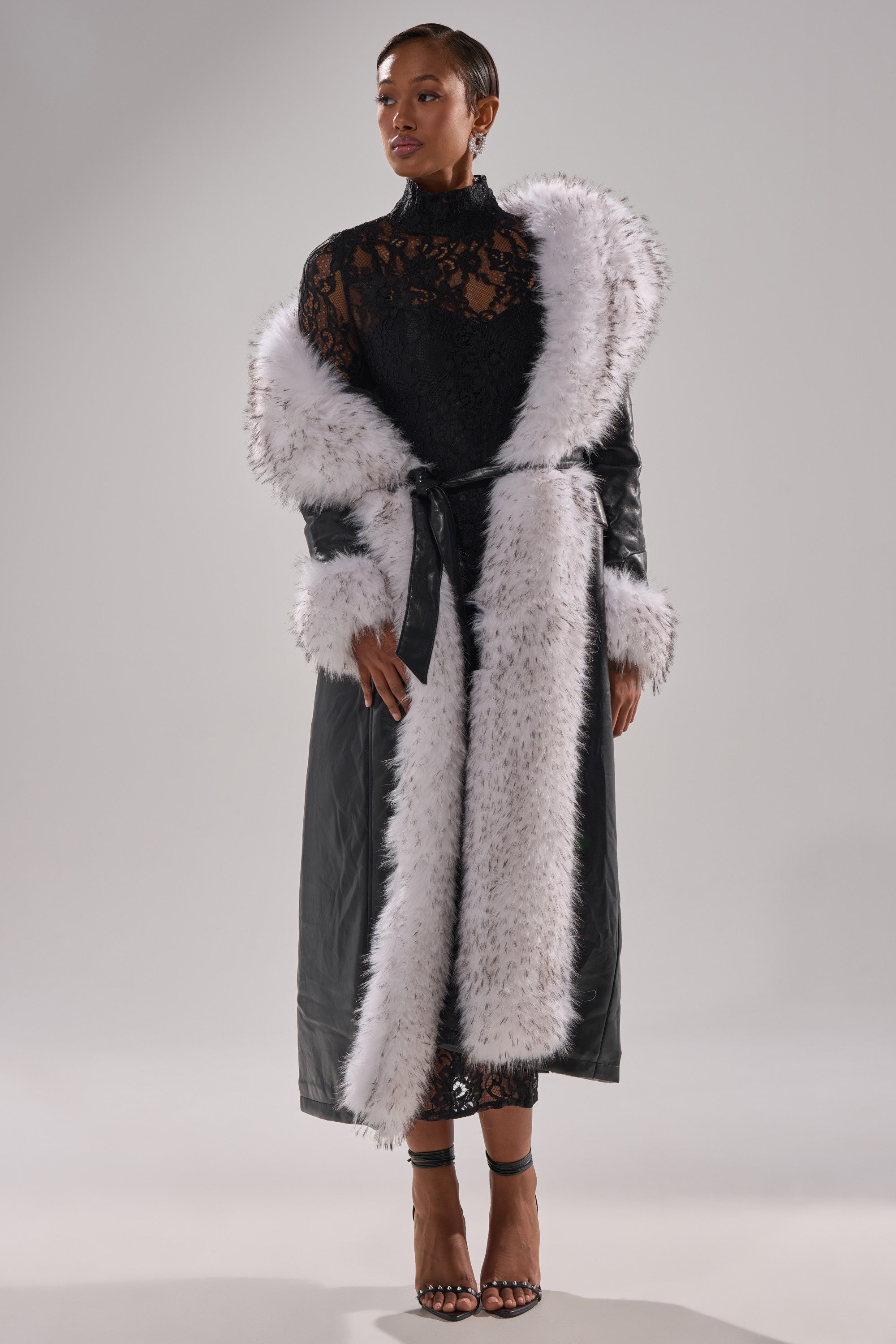 A woman stands confidently in high-heeled sandals, wearing a long black dress and the FIONA BUTTER LUXE FUR TRIM TRENCH against a plain light background.