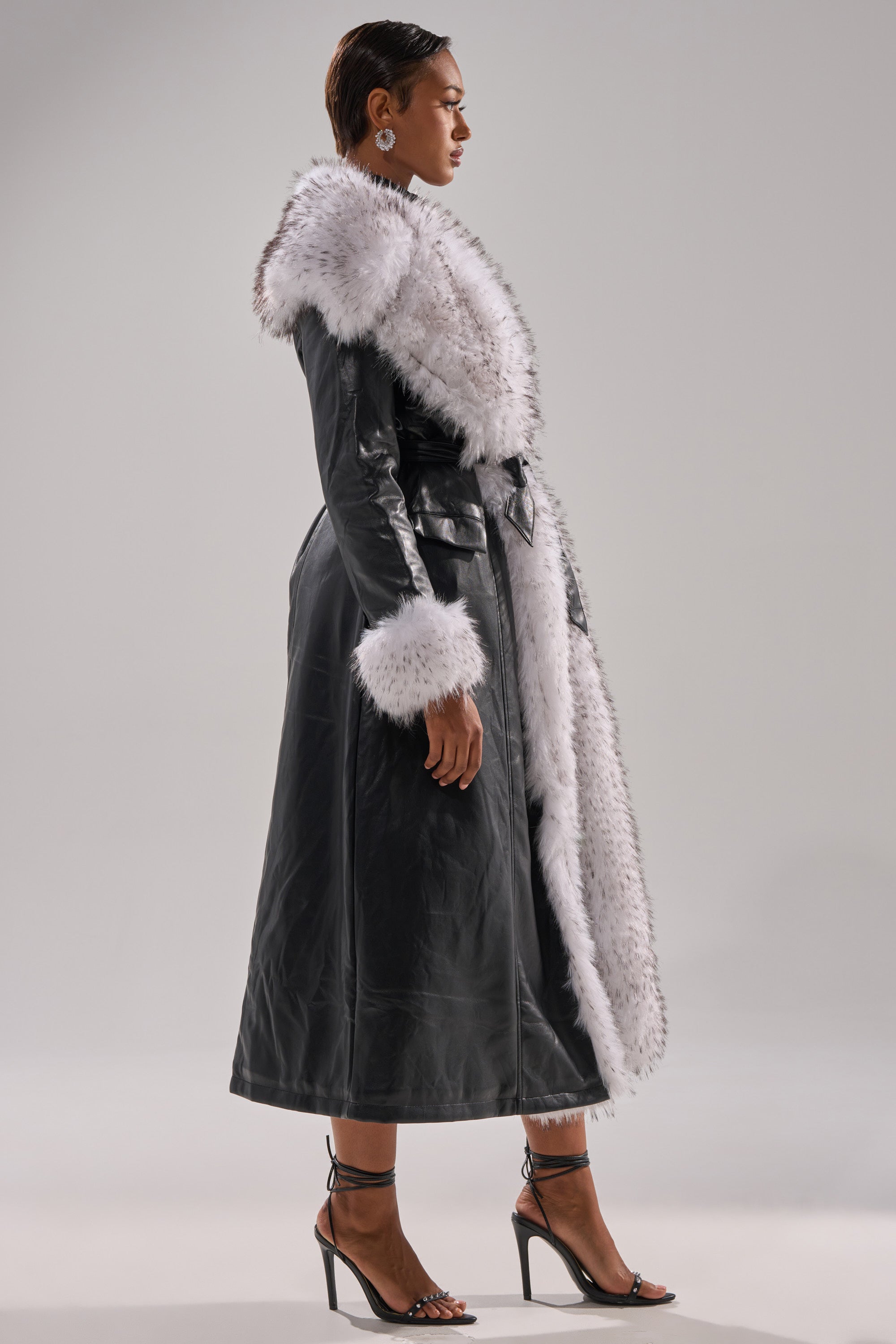 A woman stands in profile wearing the FIONA BUTTER LUXE FUR TRIM TRENCH, a black faux leather longline coat with white fur trim on the collar, cuffs, and front. She pairs it with black heels and hoop earrings against a plain light gray backdrop.